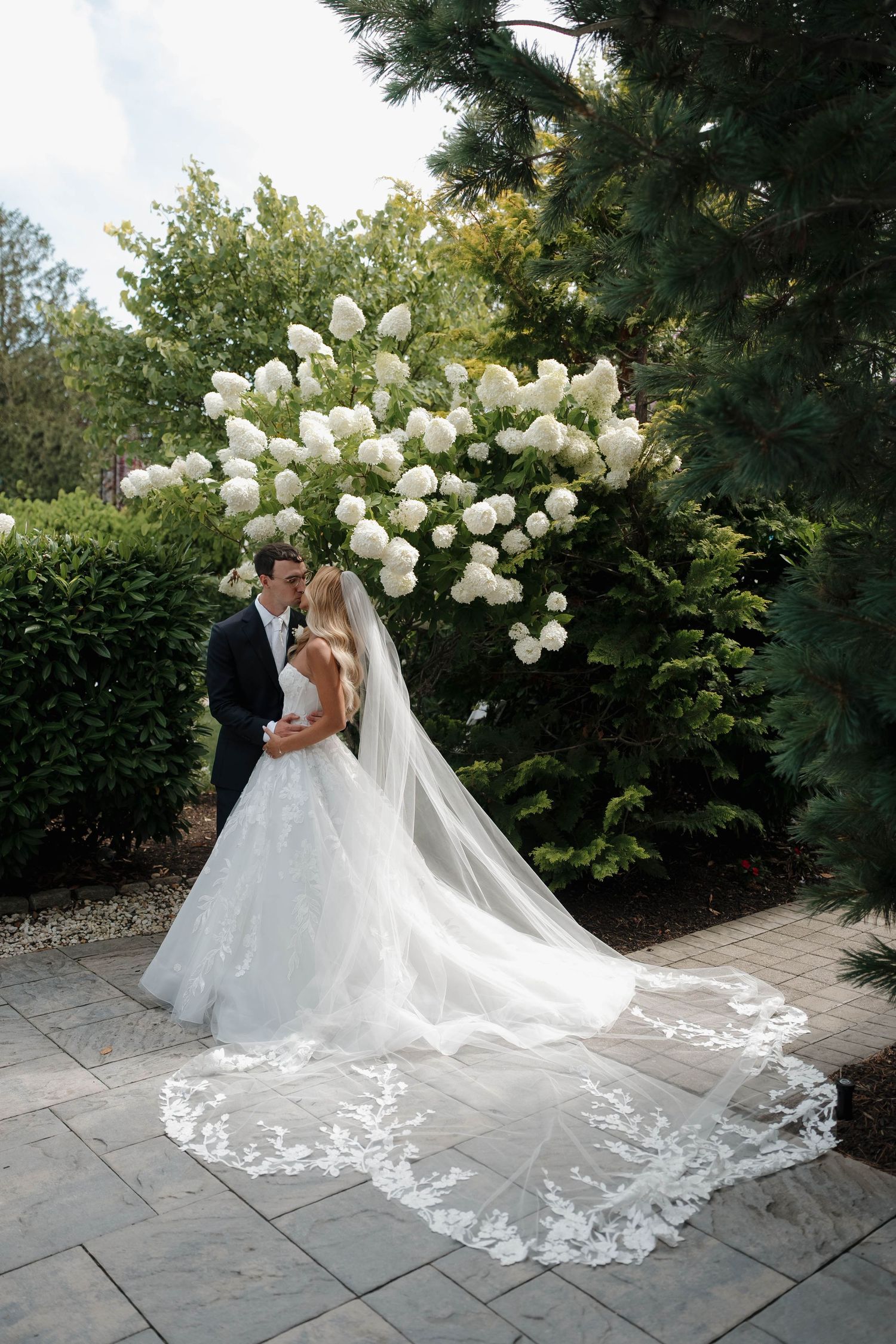 Bride and groom kissing in a garden with white hydrangeas, bride wearing a lace ball gown with cathedral veil.