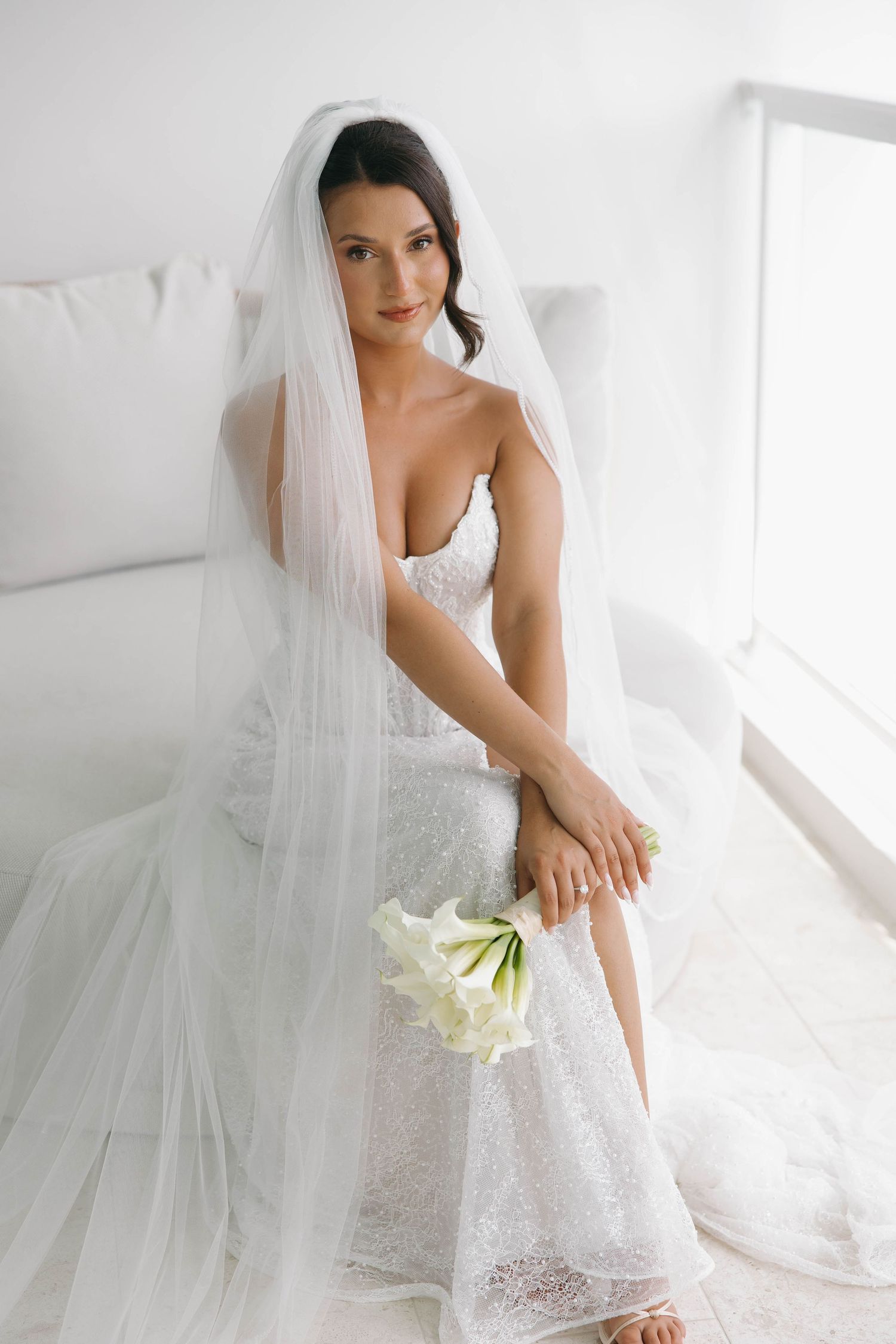 Elegant bride in white lace gown and veil sits gracefully holding white calla lilies in a bright, airy room.