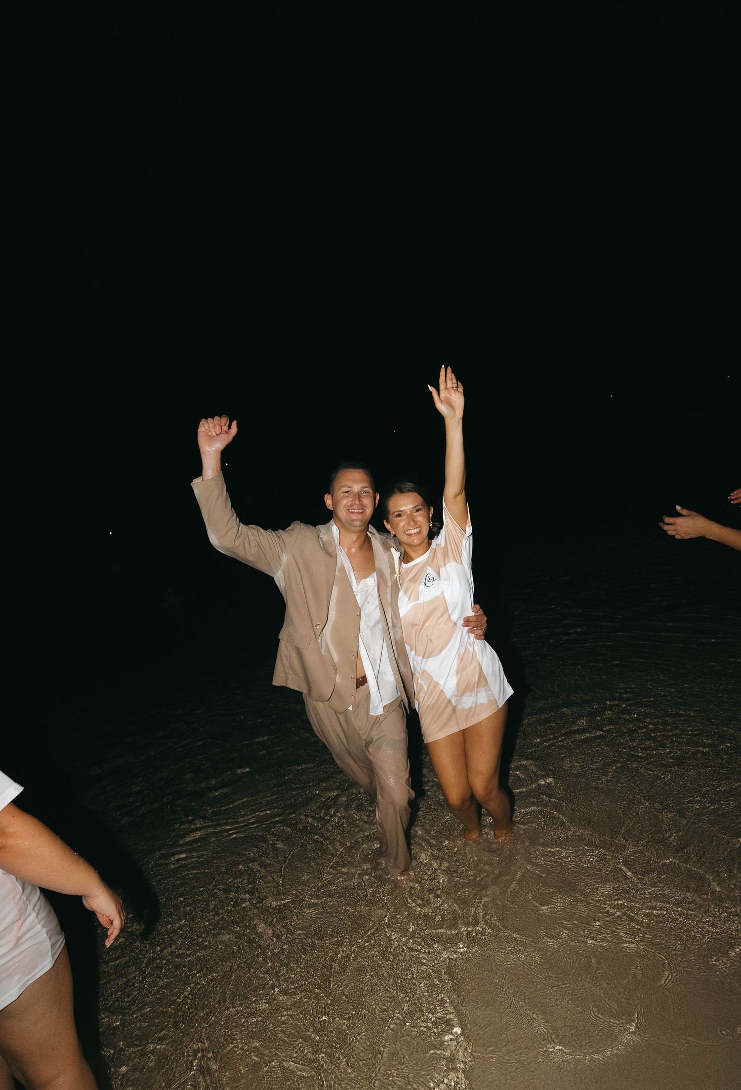 Joyful newlywed couple celebrates in ocean waves at night, wearing wedding attire with hands raised in excitement.