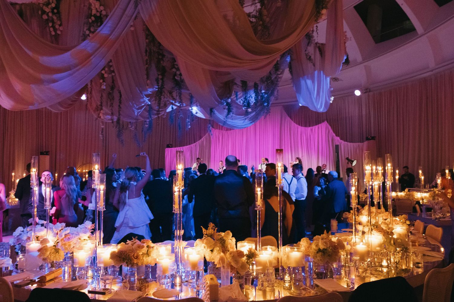 Elegant wedding reception with pink uplighting, draped ceiling, and candlelit tables.