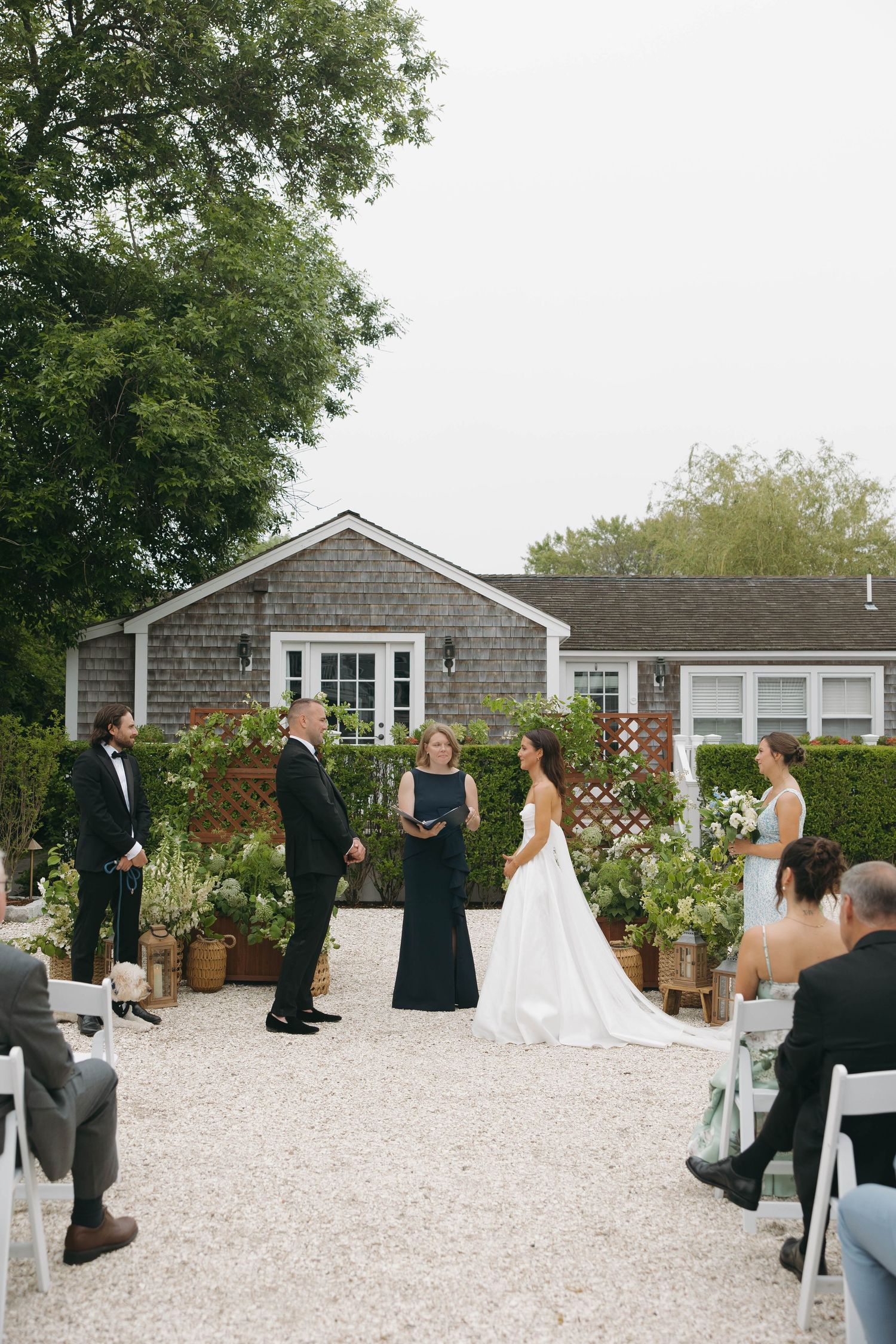 Outdoor wedding ceremony setup with white chairs and gravel ground in front of a gray shingled house.