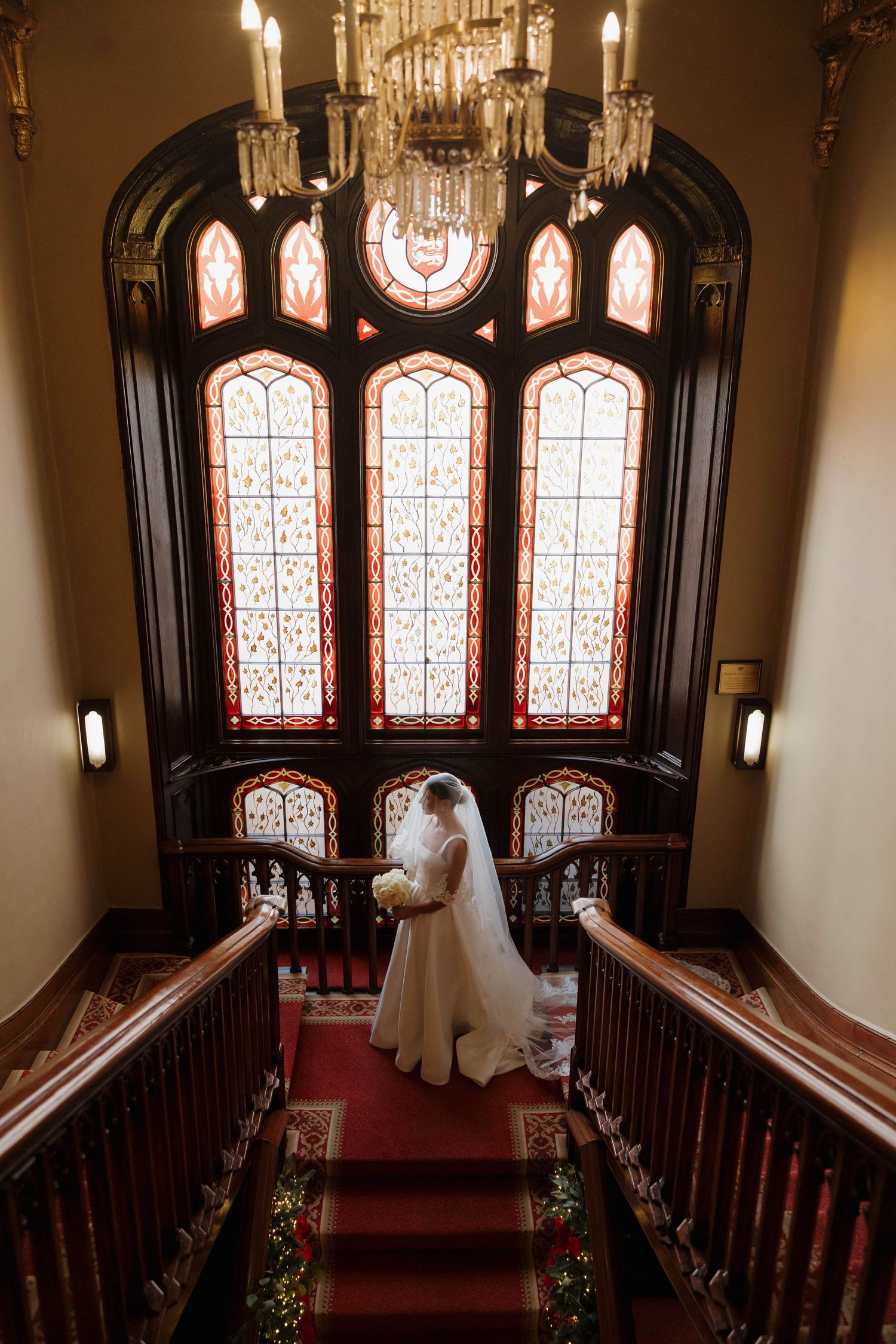 Dramatic bridal portrait on grand staircase with ornate stained glass windows and chandelier.