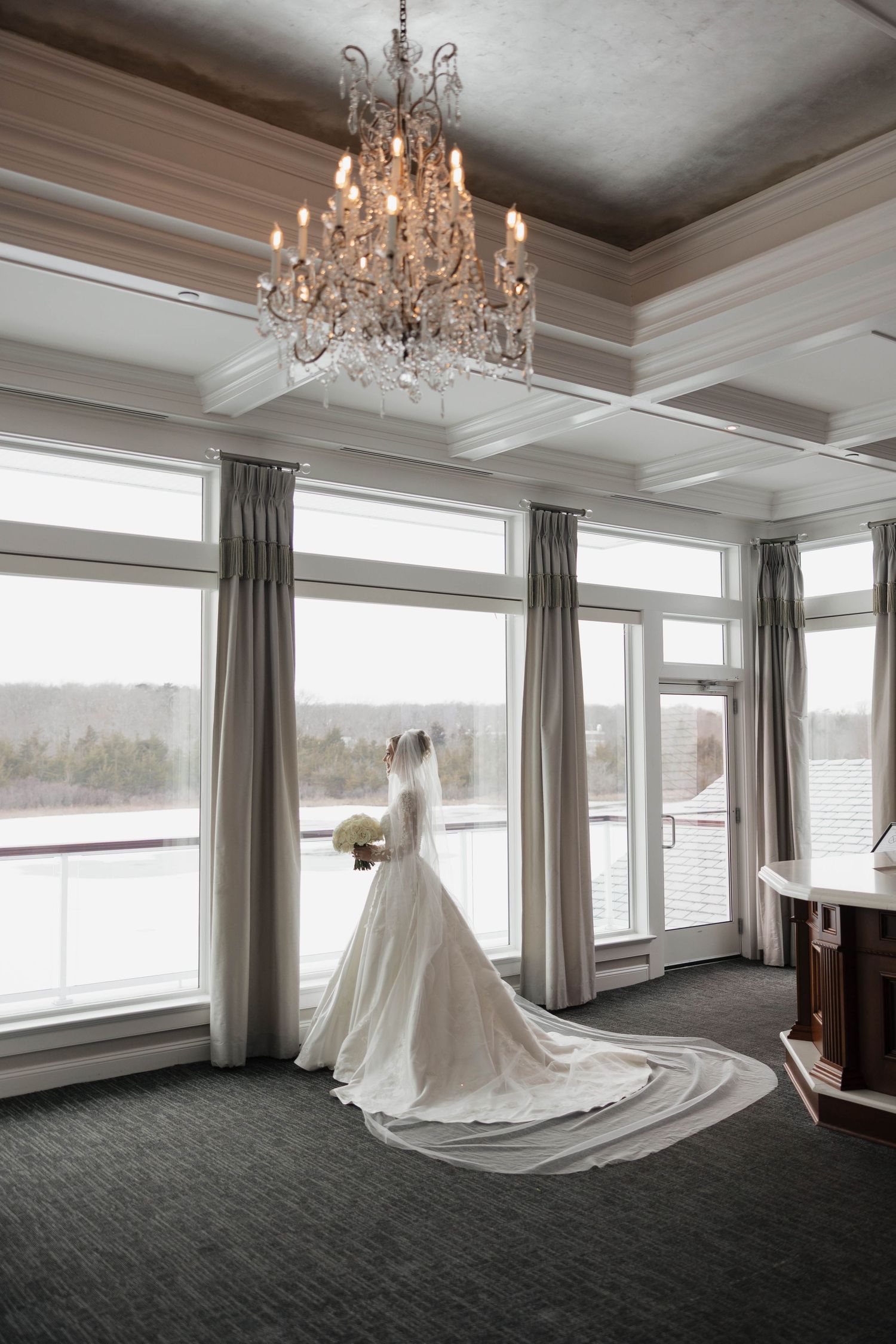 Sunlit room with chandelier and wedding dress displayed against large windows overlooking winter landscape.