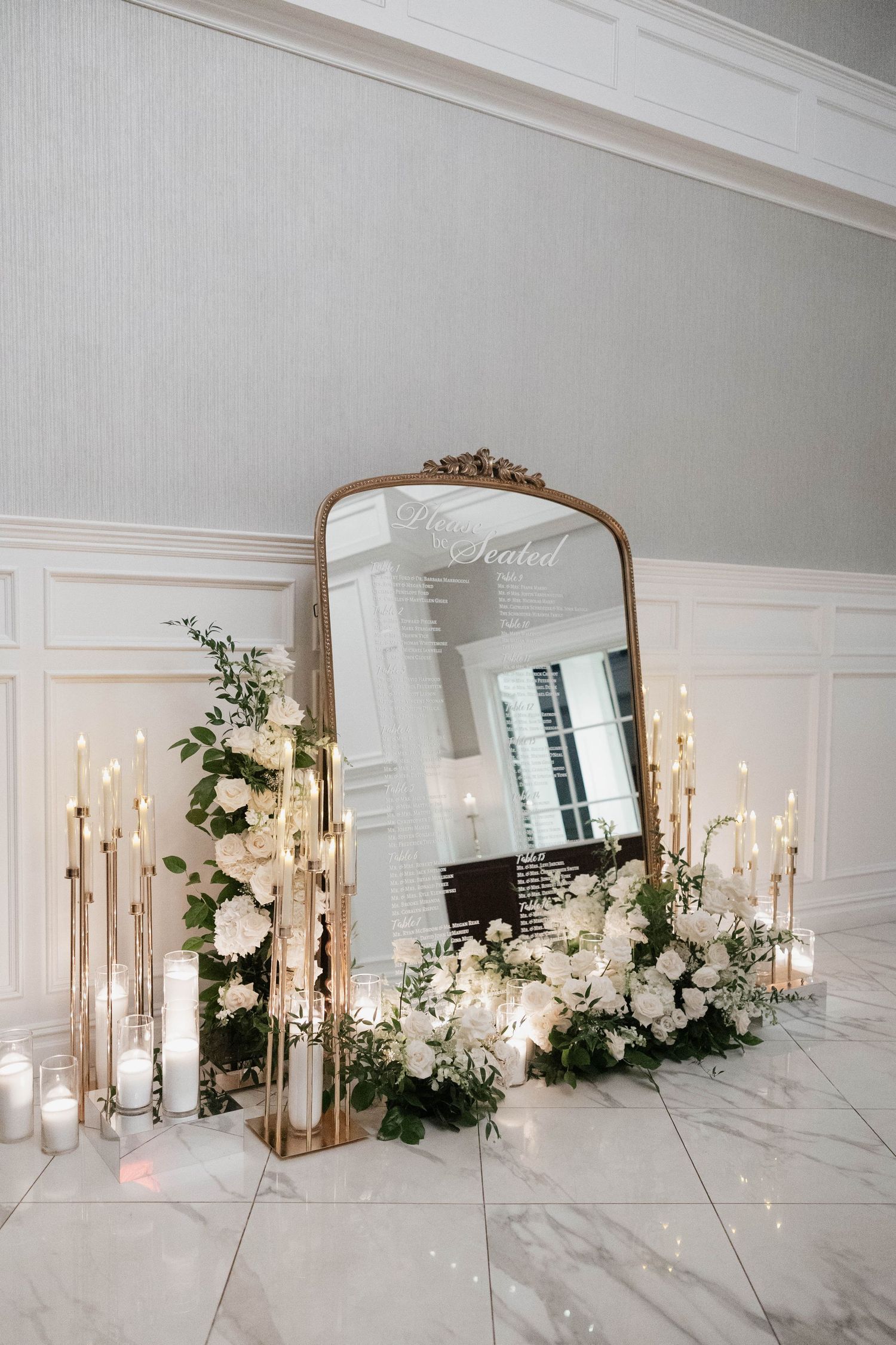 Elegant mirror display with white flowers and tall candles on marble floor.