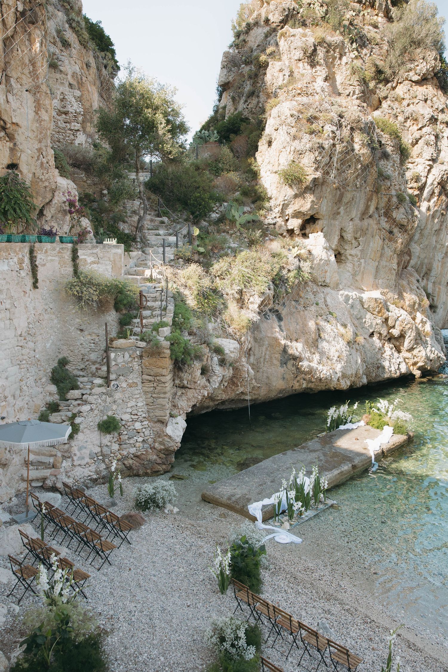Ancient stone grotto overlooking a pristine Mediterranean beach with ceremony seating arranged below.