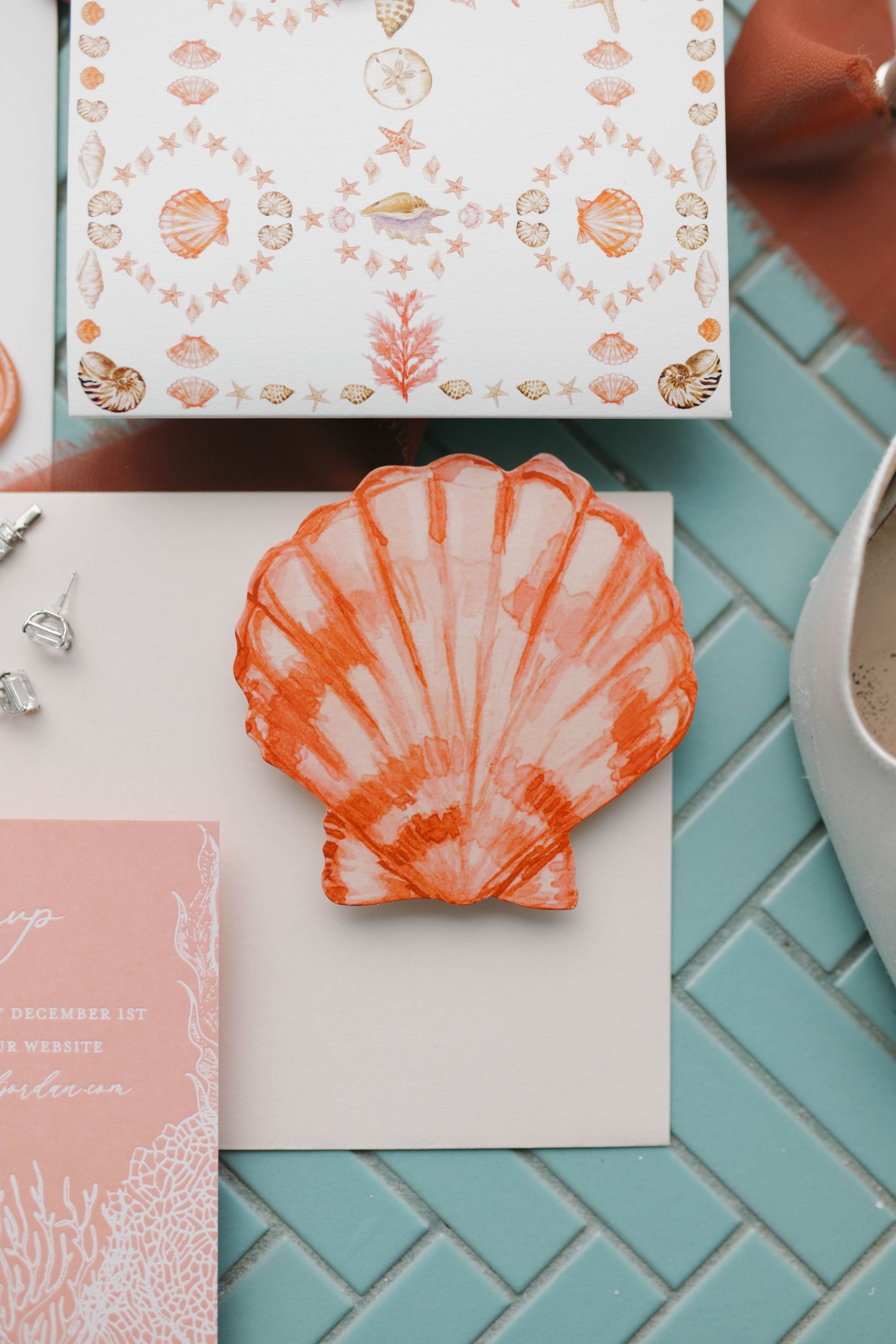 Coral-colored seashell decoration with wedding stationery on turquoise tile surface.