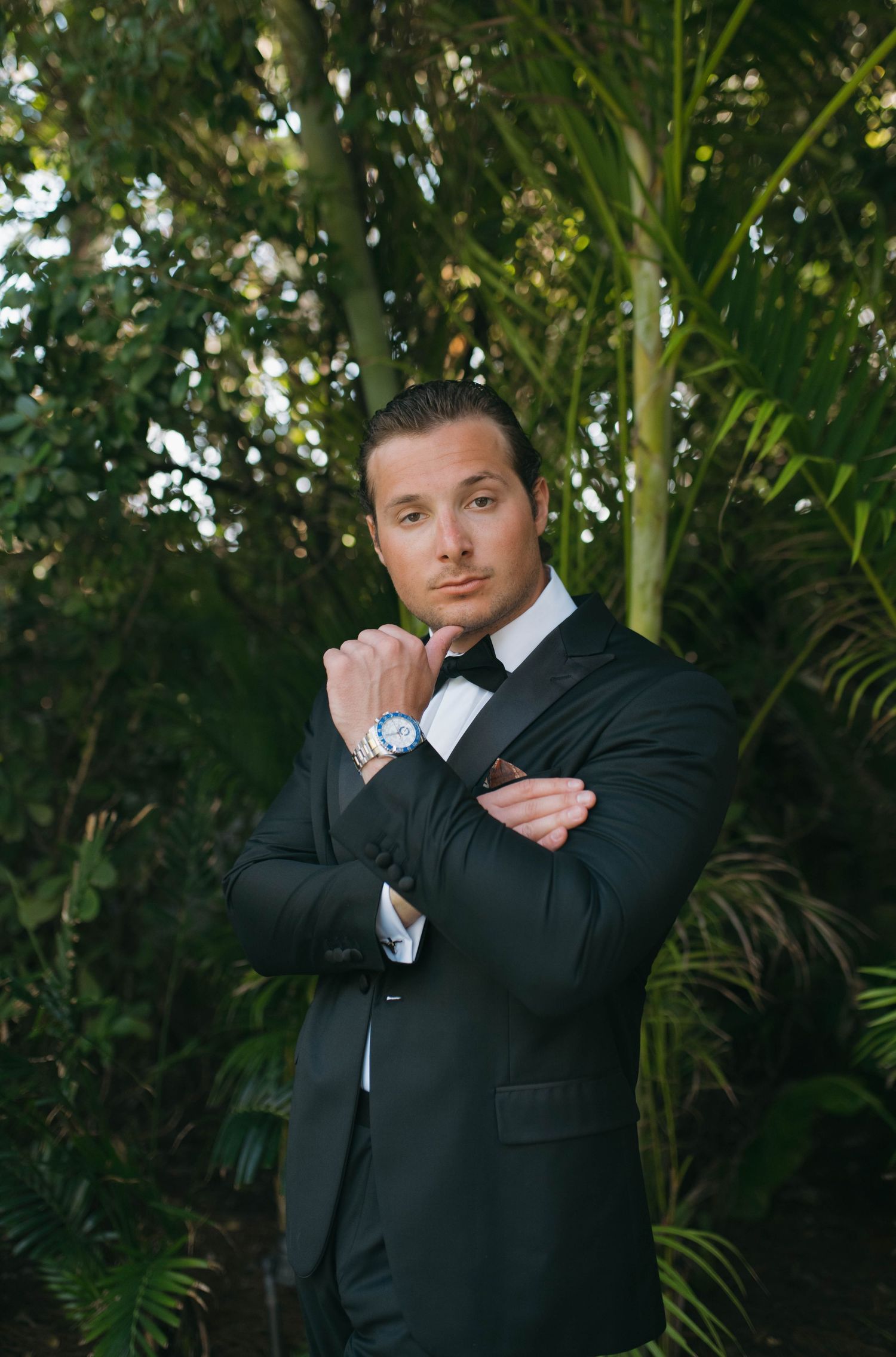 Groom in black tuxedo poses playfully in tropical garden setting.