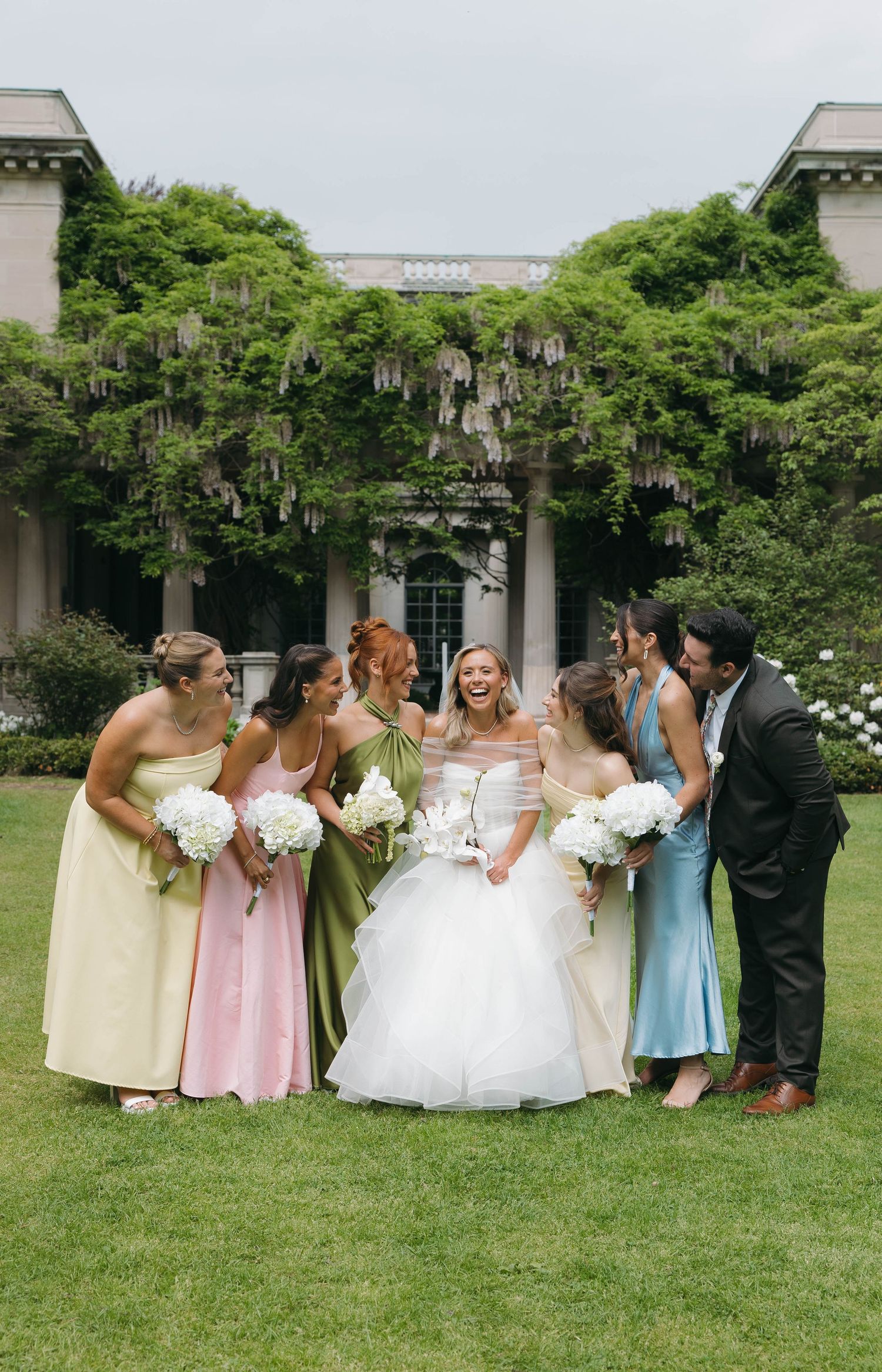 Wedding party in pastel dresses posed together on grass in front of columned building.