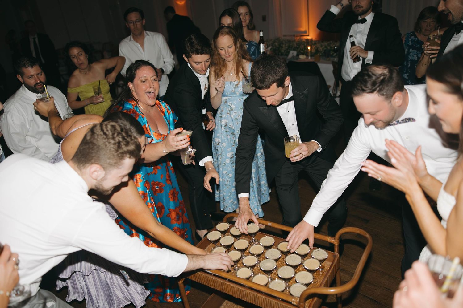 Party guests reach for shots from a wooden serving tray at an indoor celebration.