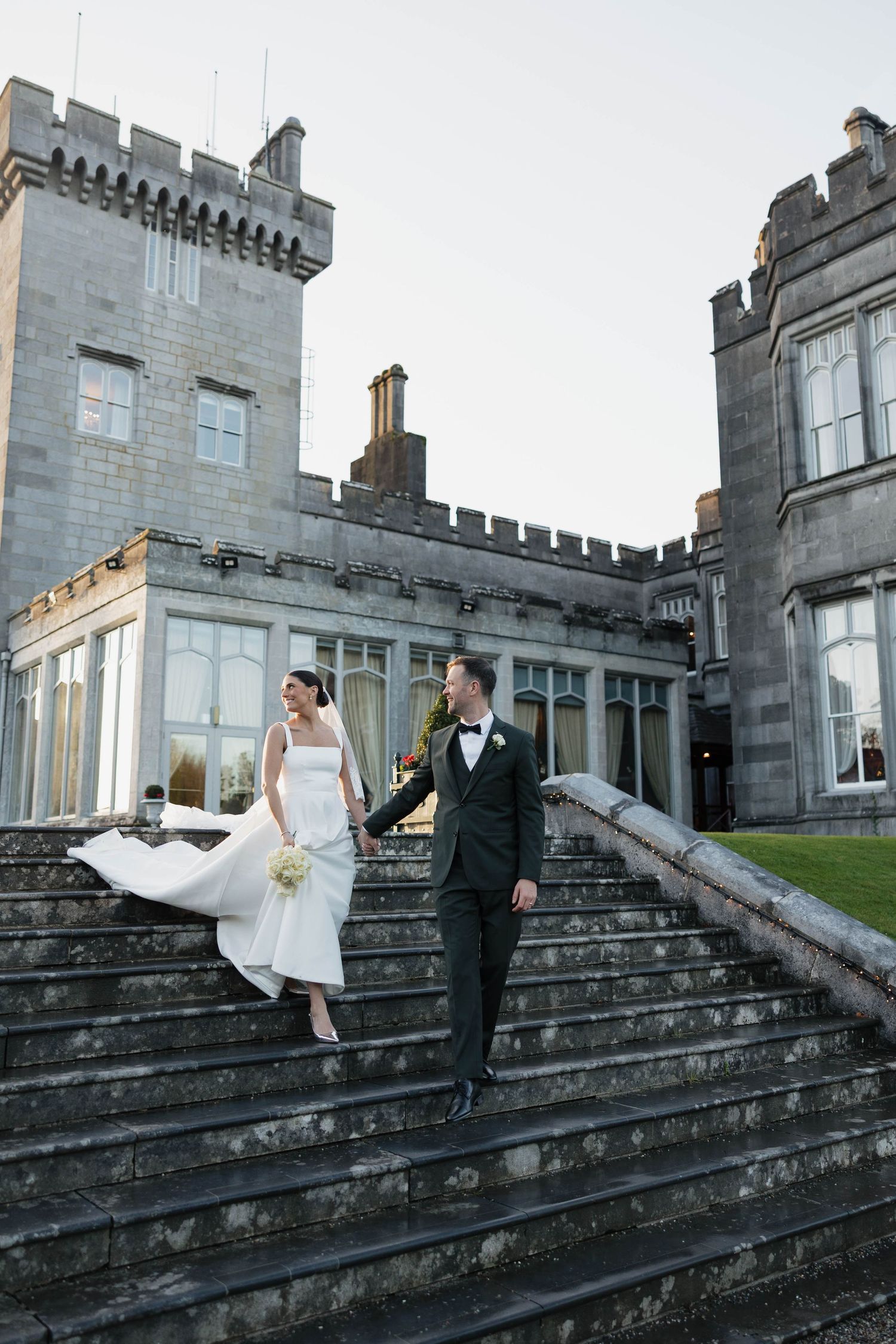 Wedding couple descends grand stone steps of historic castle.