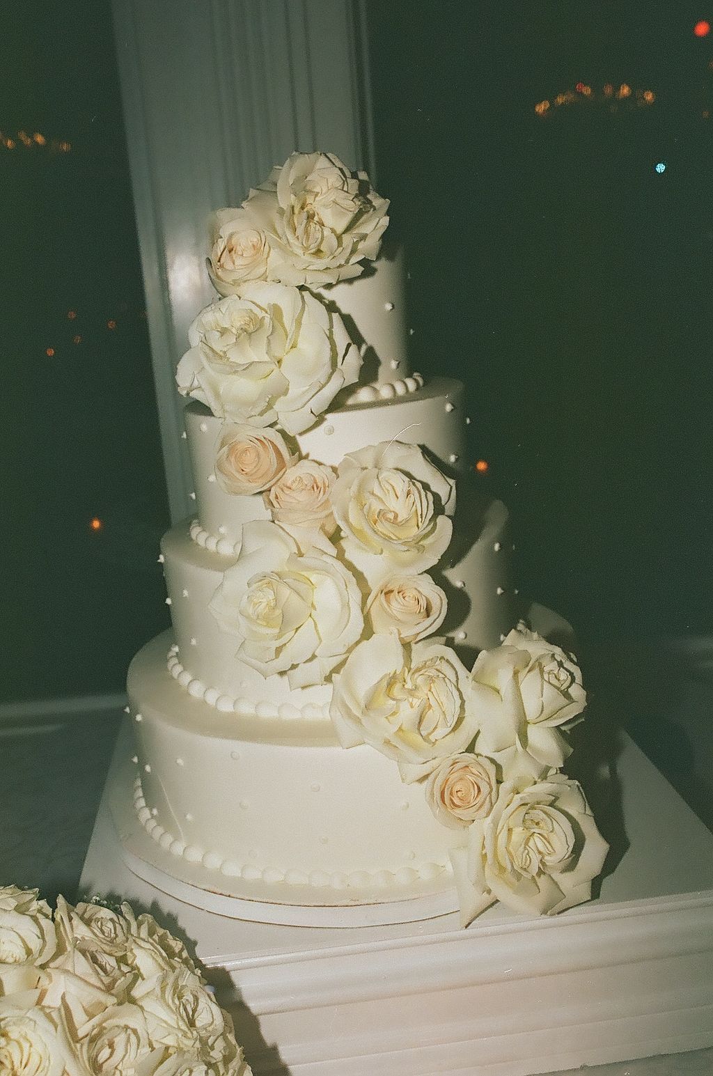 Elegant white wedding cake decorated with cascading cream roses and delicate pearl details against a dark background.