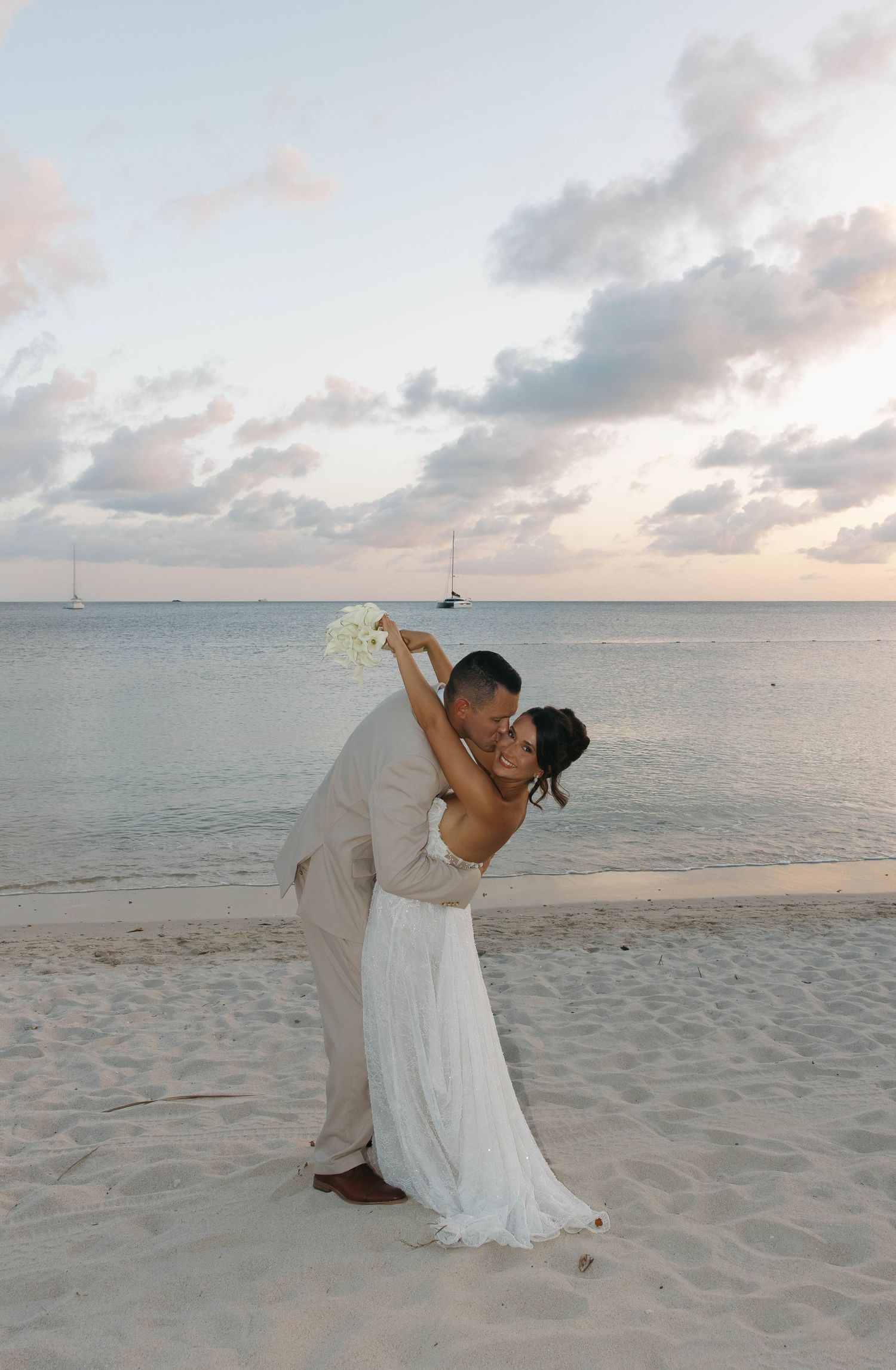 Bride and groom share romantic dip kiss on sandy beach at sunset with ocean and sailboat backdrop.