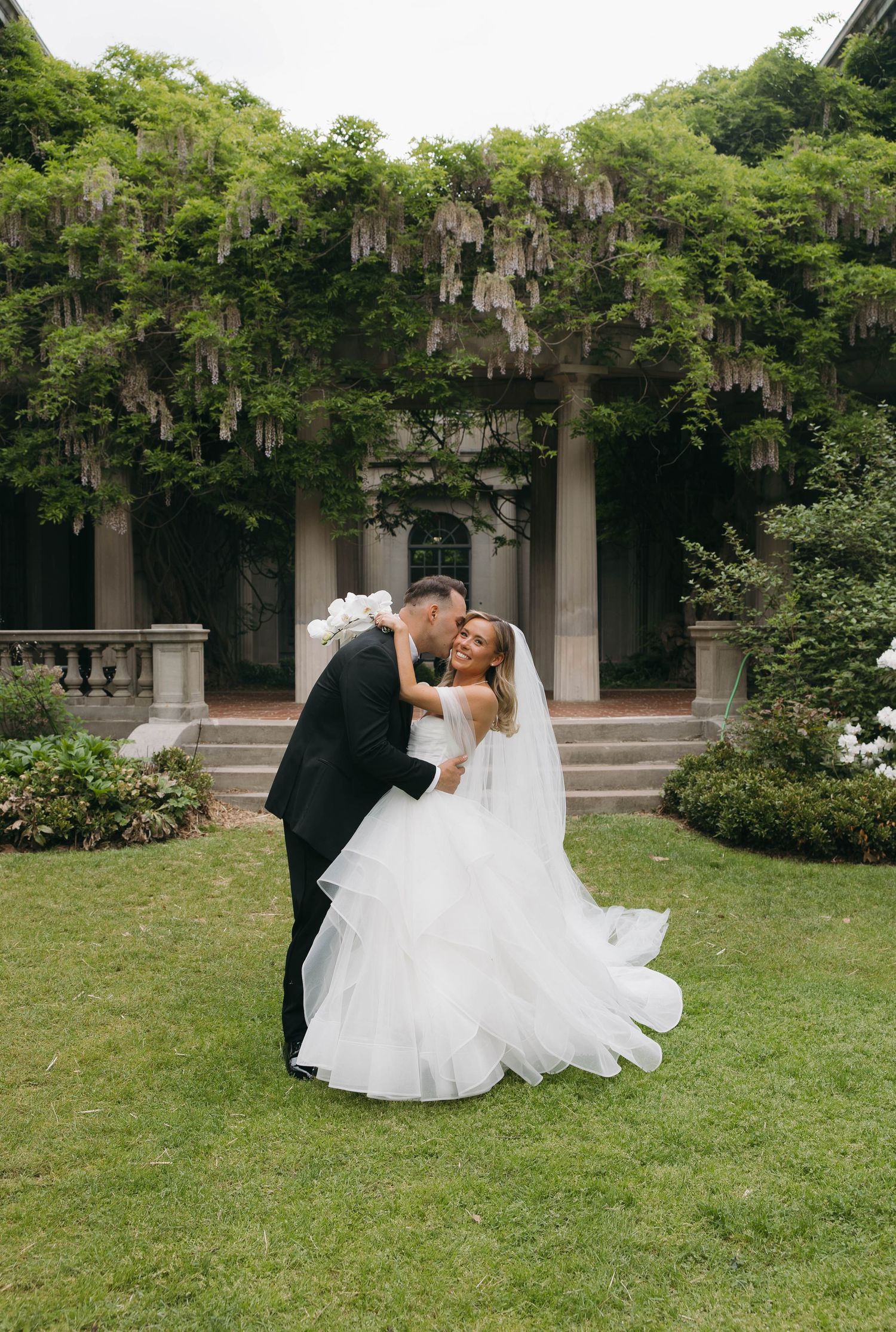 Romantic wedding couple embracing on manicured garden grounds with classical architecture.