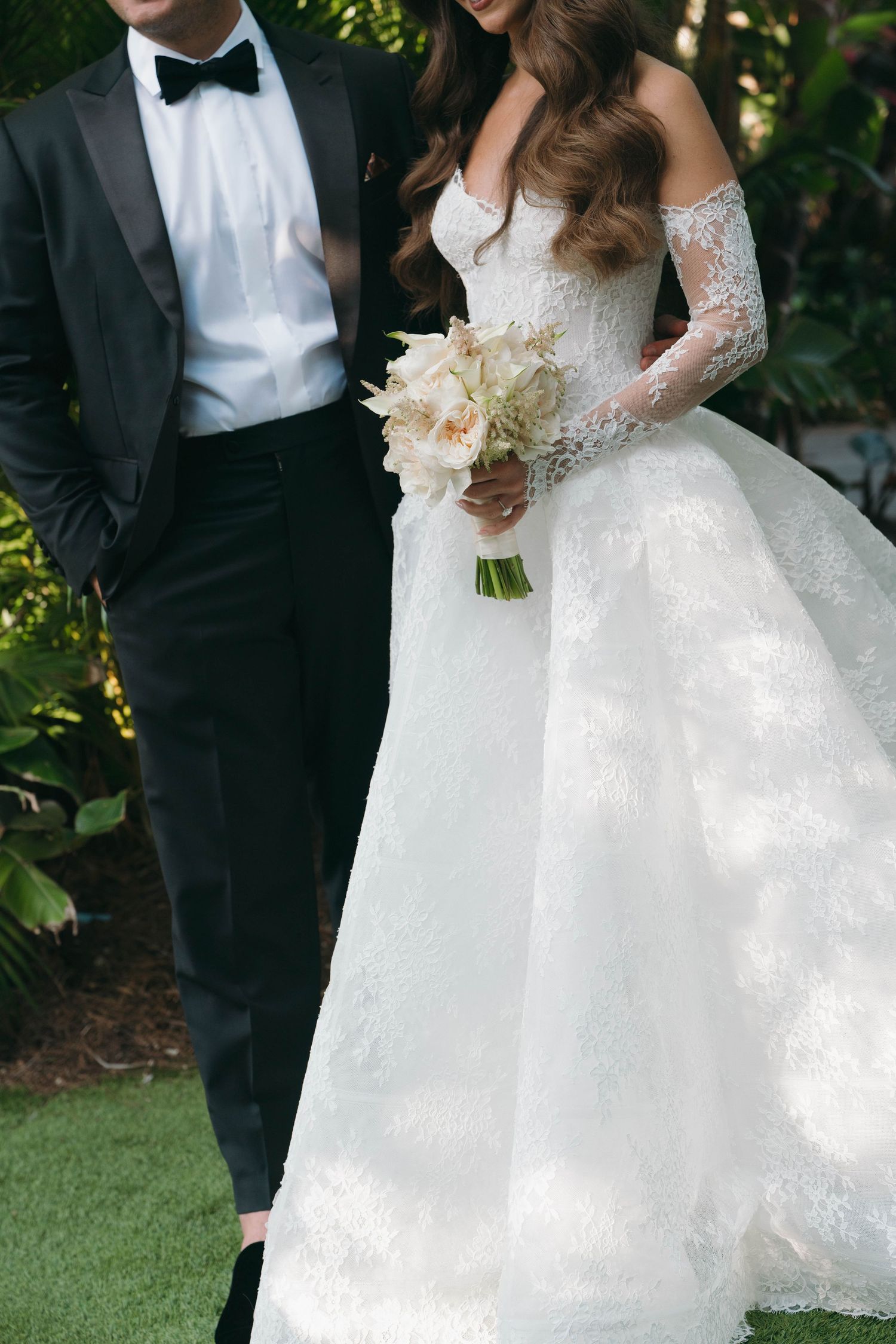 Wedding couple in formal attire posing outdoors with white floral bouquet.