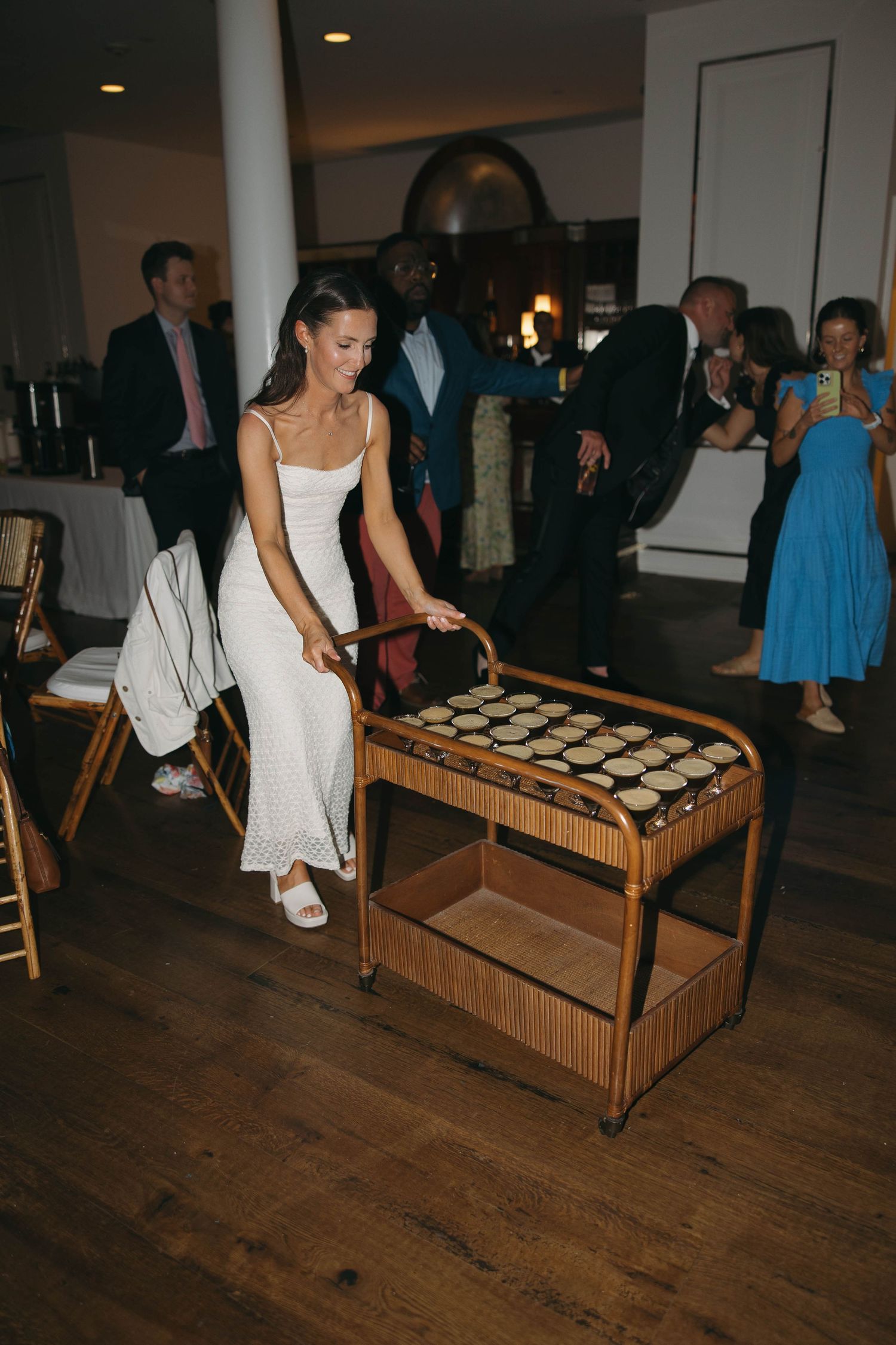Wedding guests gather around a wooden tray filled with shot glasses at a reception party.