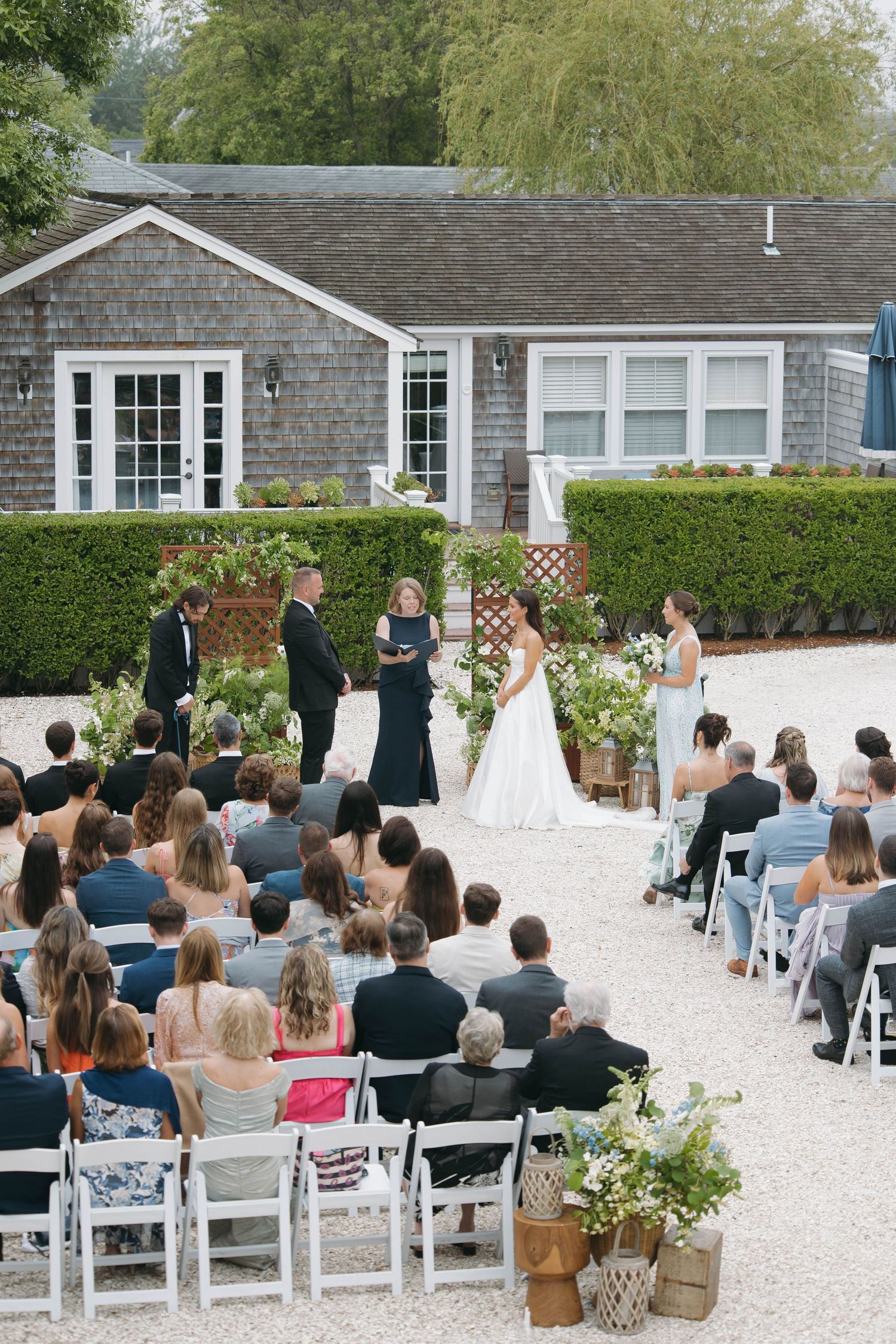 Wedding guests seated in white chairs watch ceremony in front of shingle-style building.