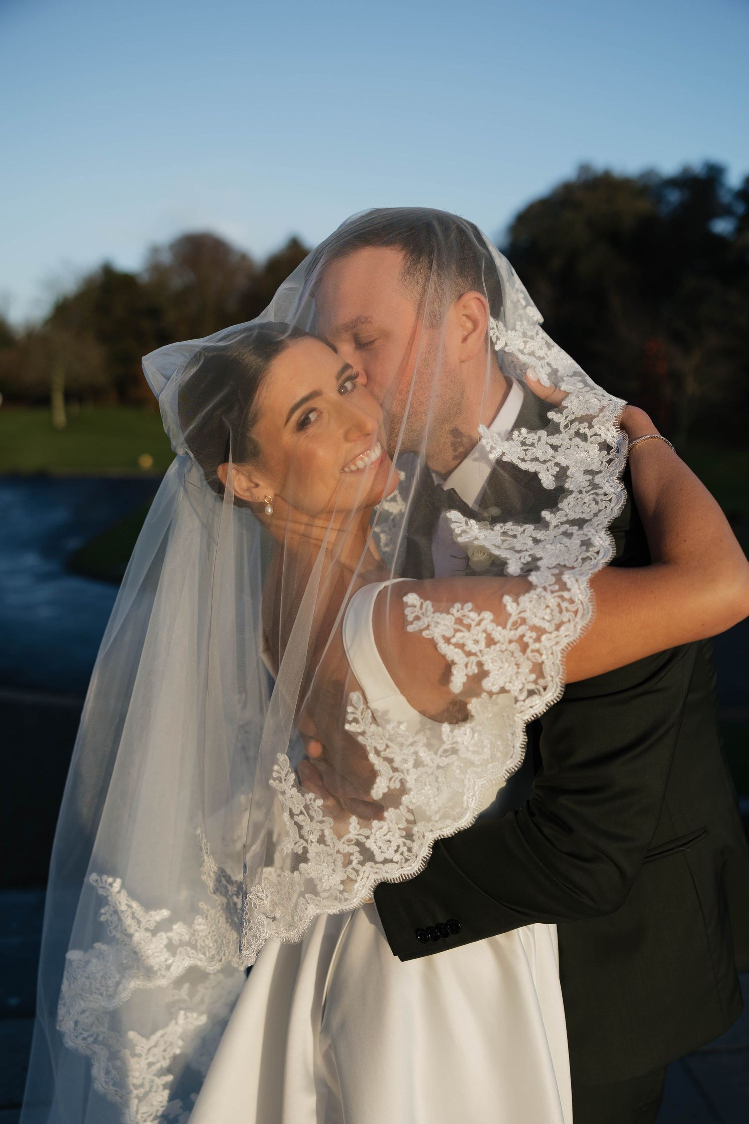 Newlyweds pose playfully with bride's lace veil in outdoor setting.