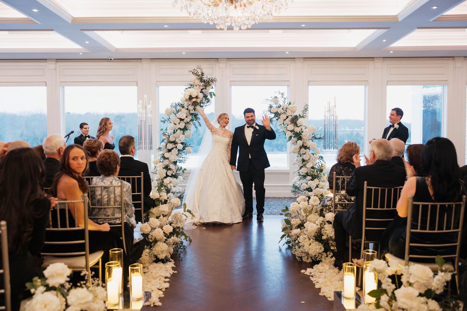 Wedding ceremony in bright venue with white floral arches and candlelit aisle.