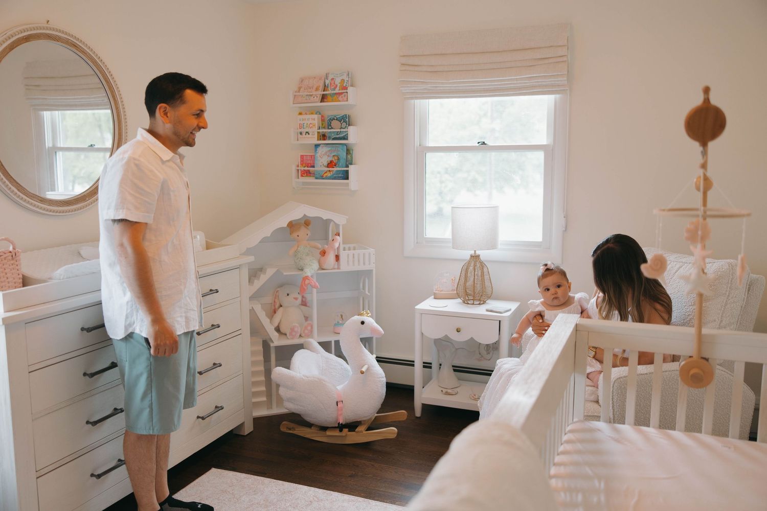 A nursery room scene with a white rocking swan and furniture including a crib, dresser, and round mirror on the wall.