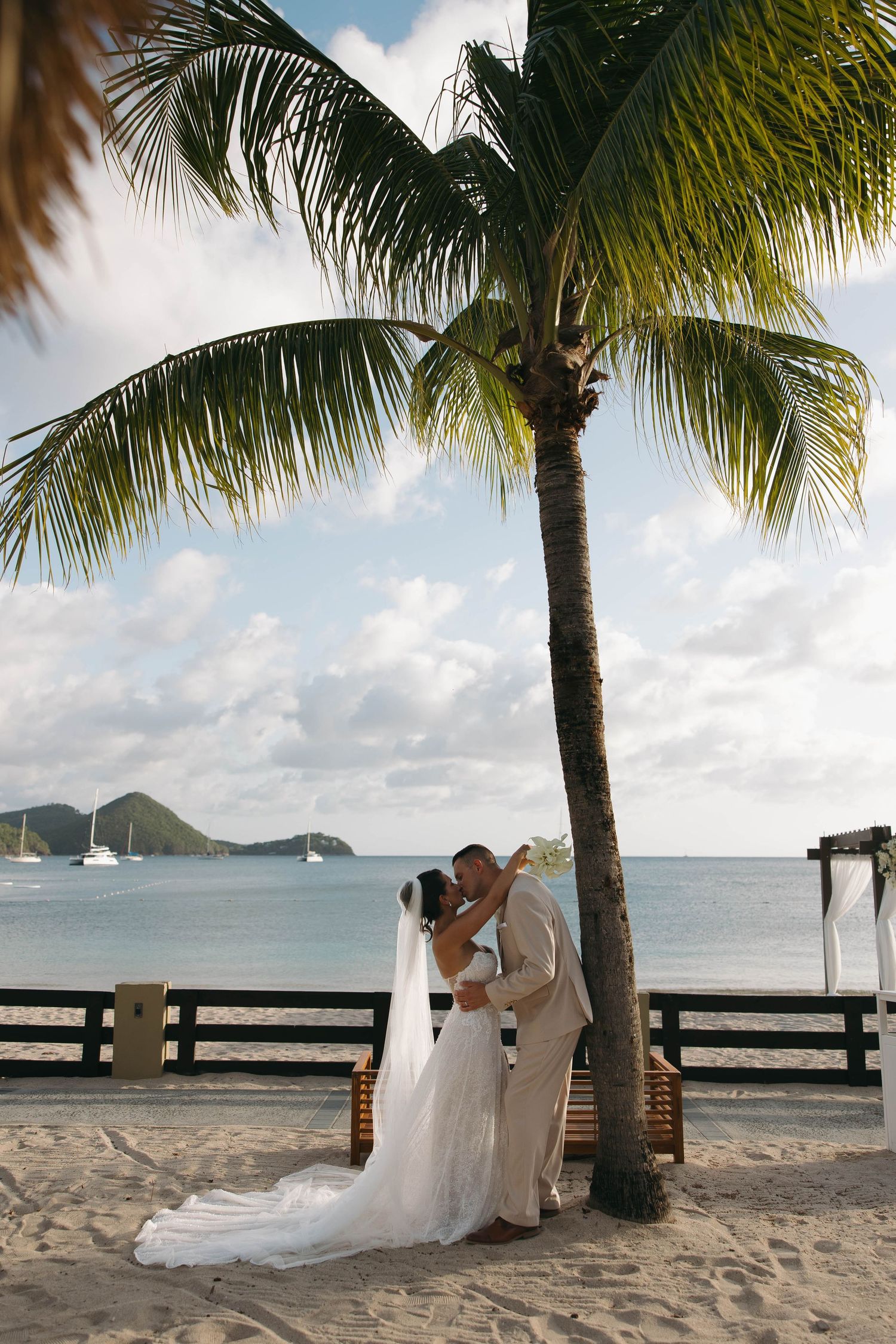 Bride and groom share a romantic kiss under a palm tree on a white sand beach during a tropical wedding ceremony.