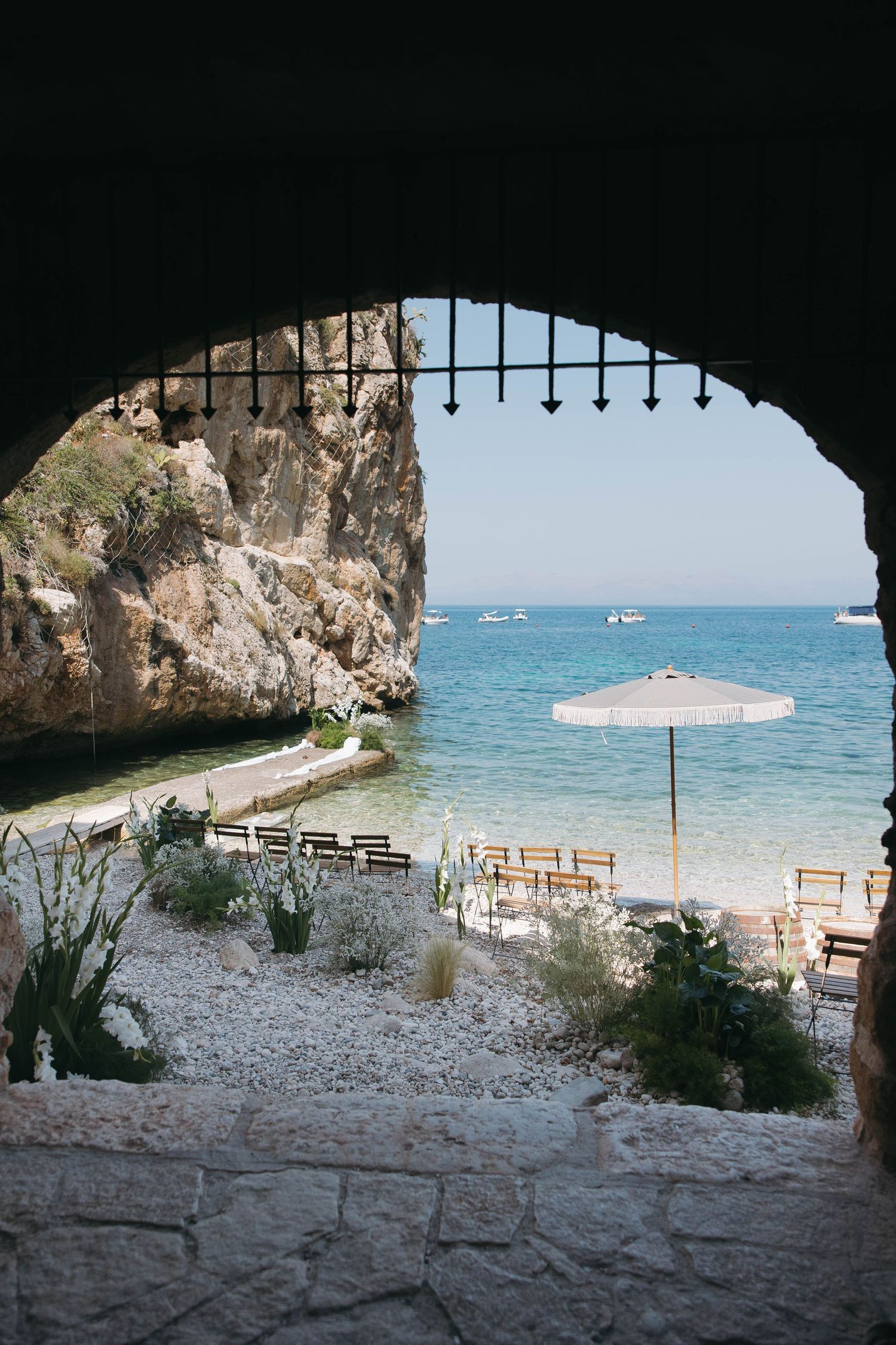 Beach wedding setup viewed through a stone archway with wooden chairs facing the turquoise sea.