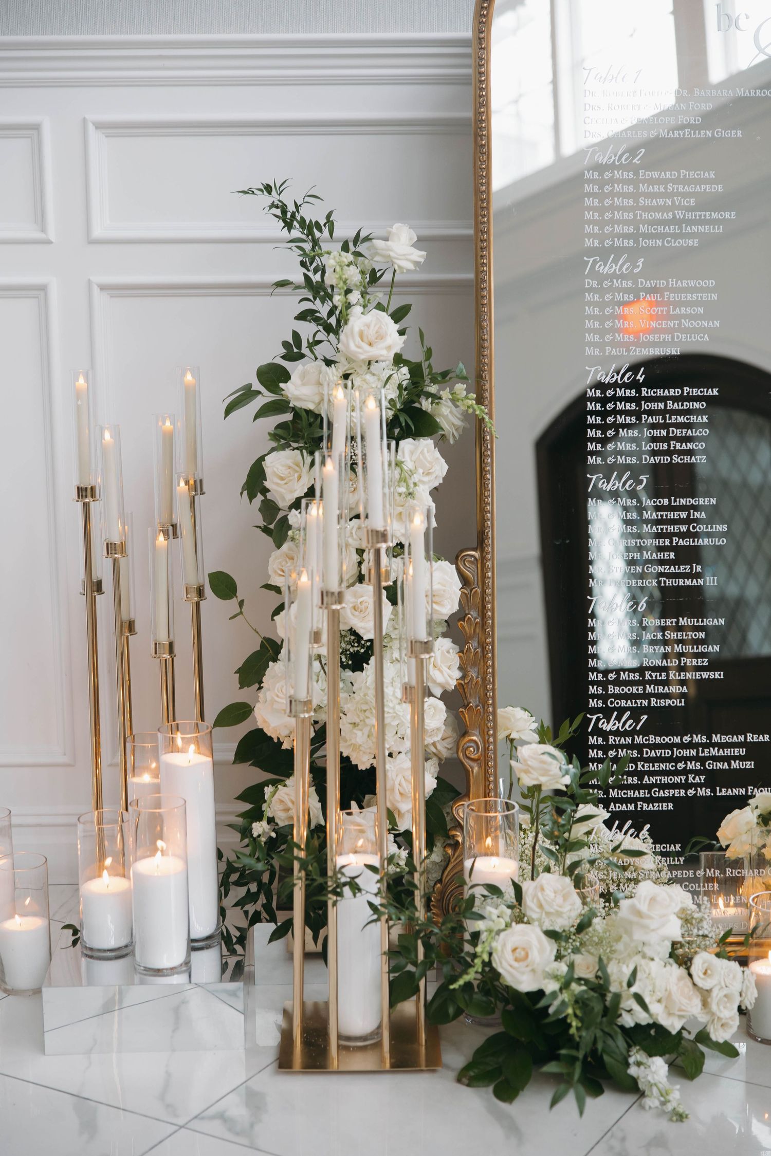 Elegant wedding decor featuring tall candles, white roses, and greenery against a white paneled wall.