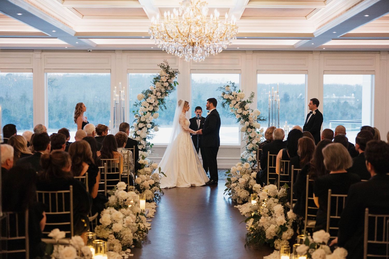 Elegant wedding venue with crystal chandelier and white floral arch overlooking waterfront view.