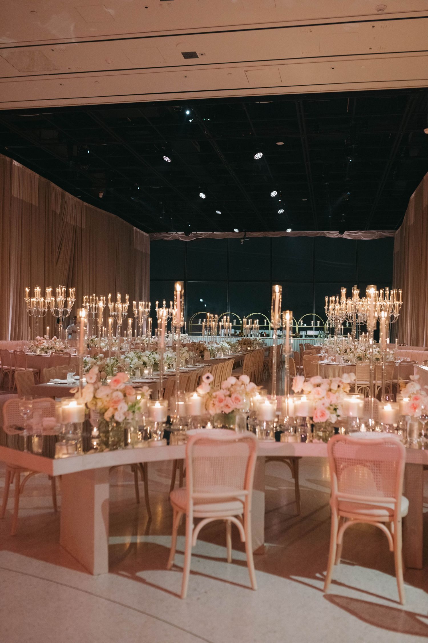 Wedding reception tables arranged in rows with crystal chandeliers and pink ambient lighting.