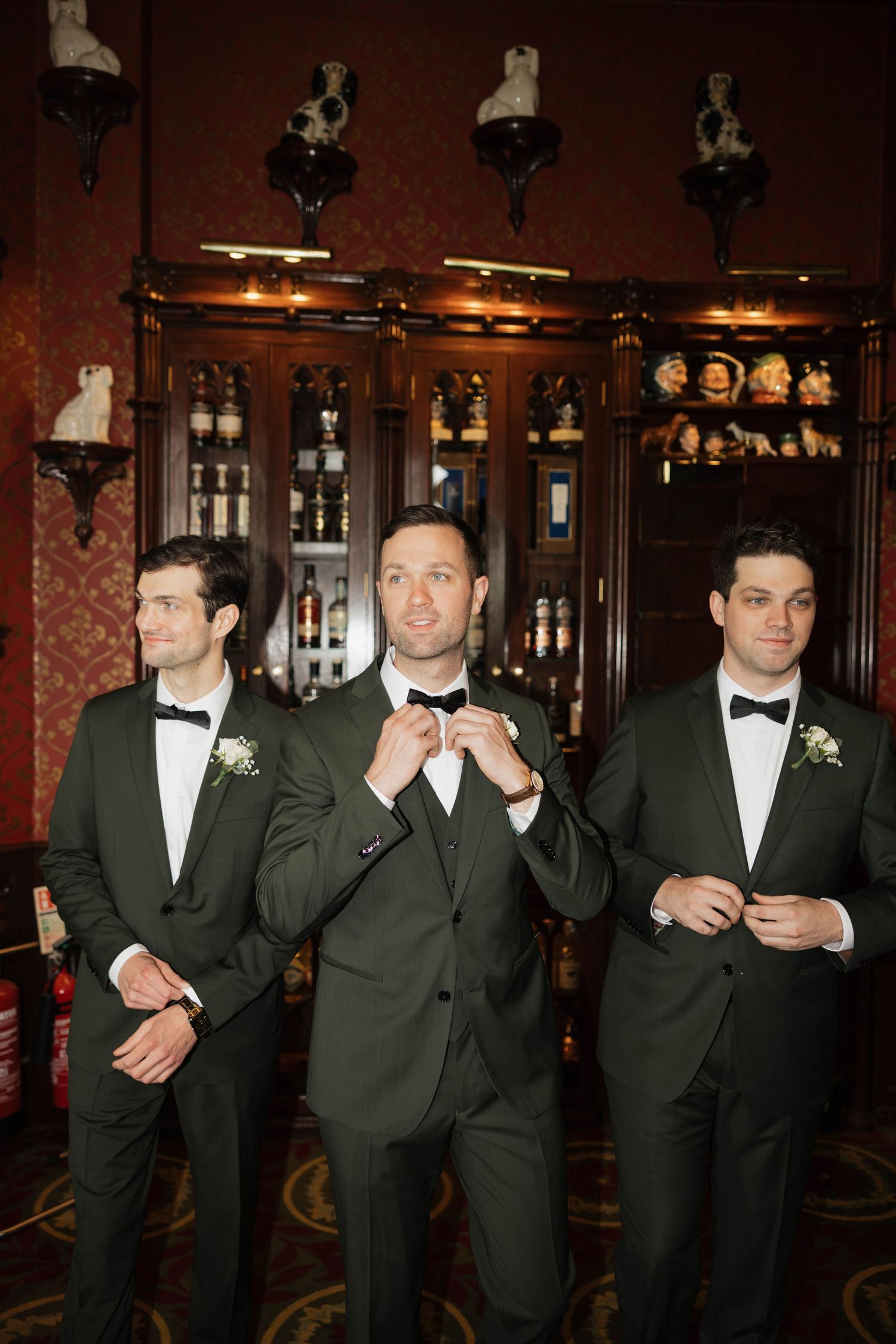 Groomsmen pose casually in black tuxedos against dark wood paneling and red walls.