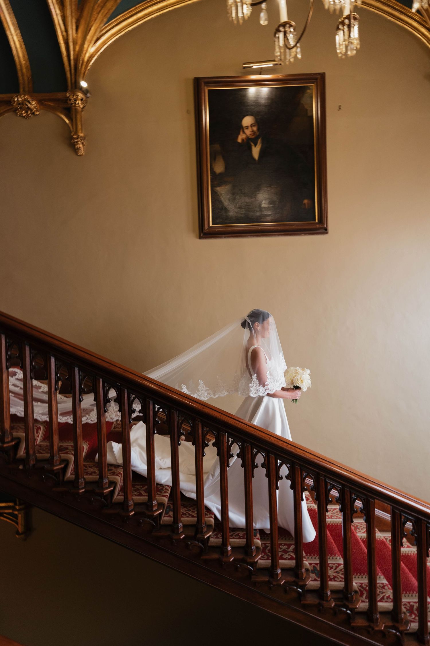Bride descending classic wooden staircase under vintage chandelier in historic venue.