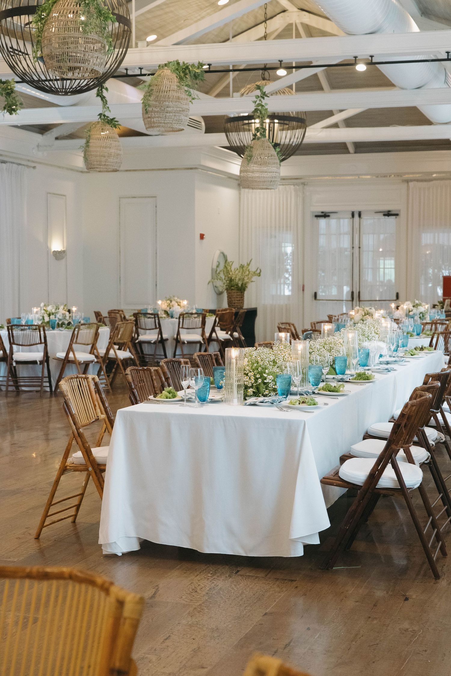 Reception hall with white-clothed round tables, wooden chairs and blue accents in a bright airy venue.