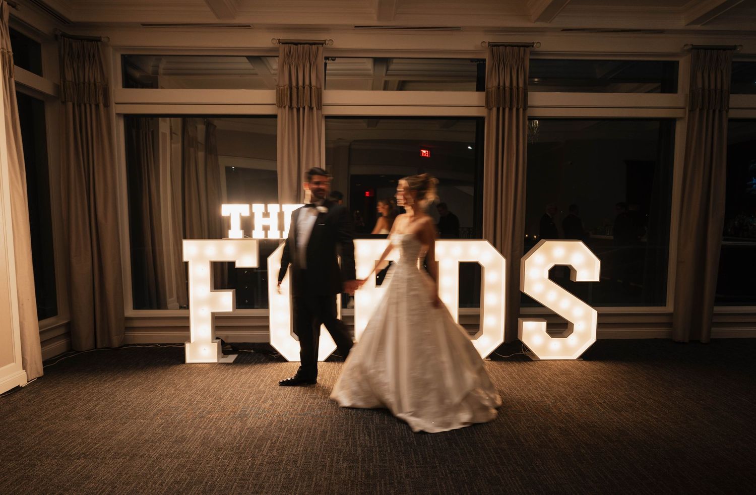 Panoramic view of a wedding reception with illuminated signage and dancing couple.