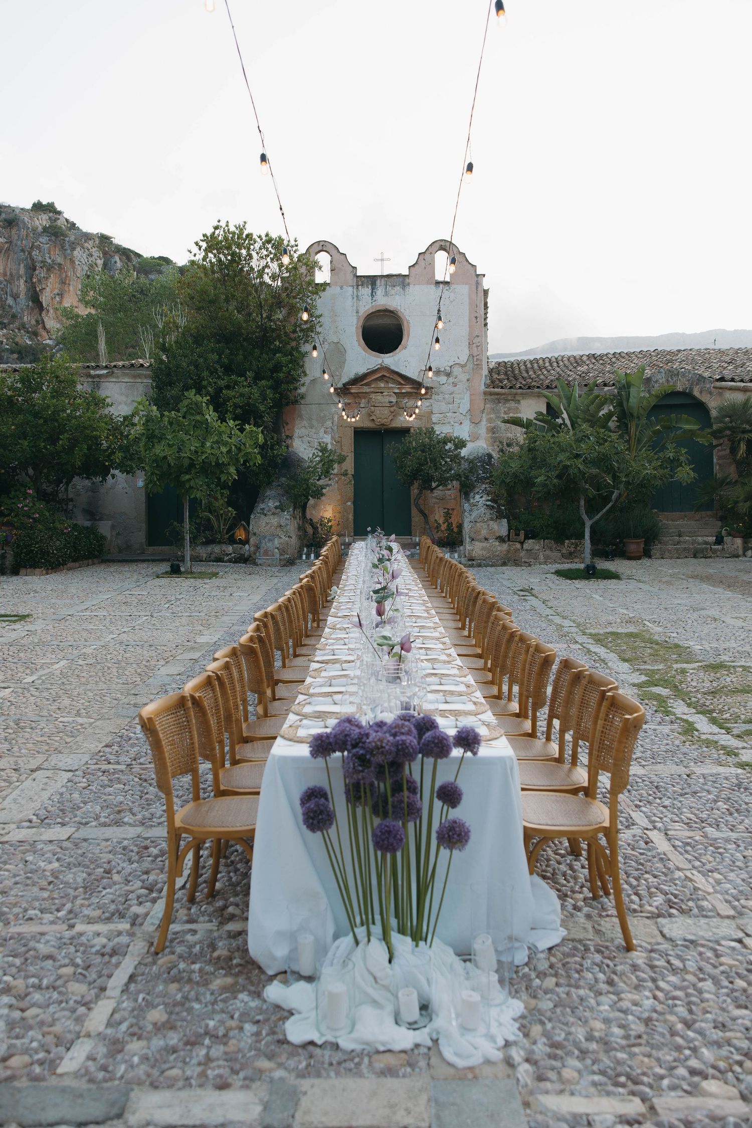 Elegant outdoor wedding reception setup with long white tables and wooden chairs in a historic courtyard.