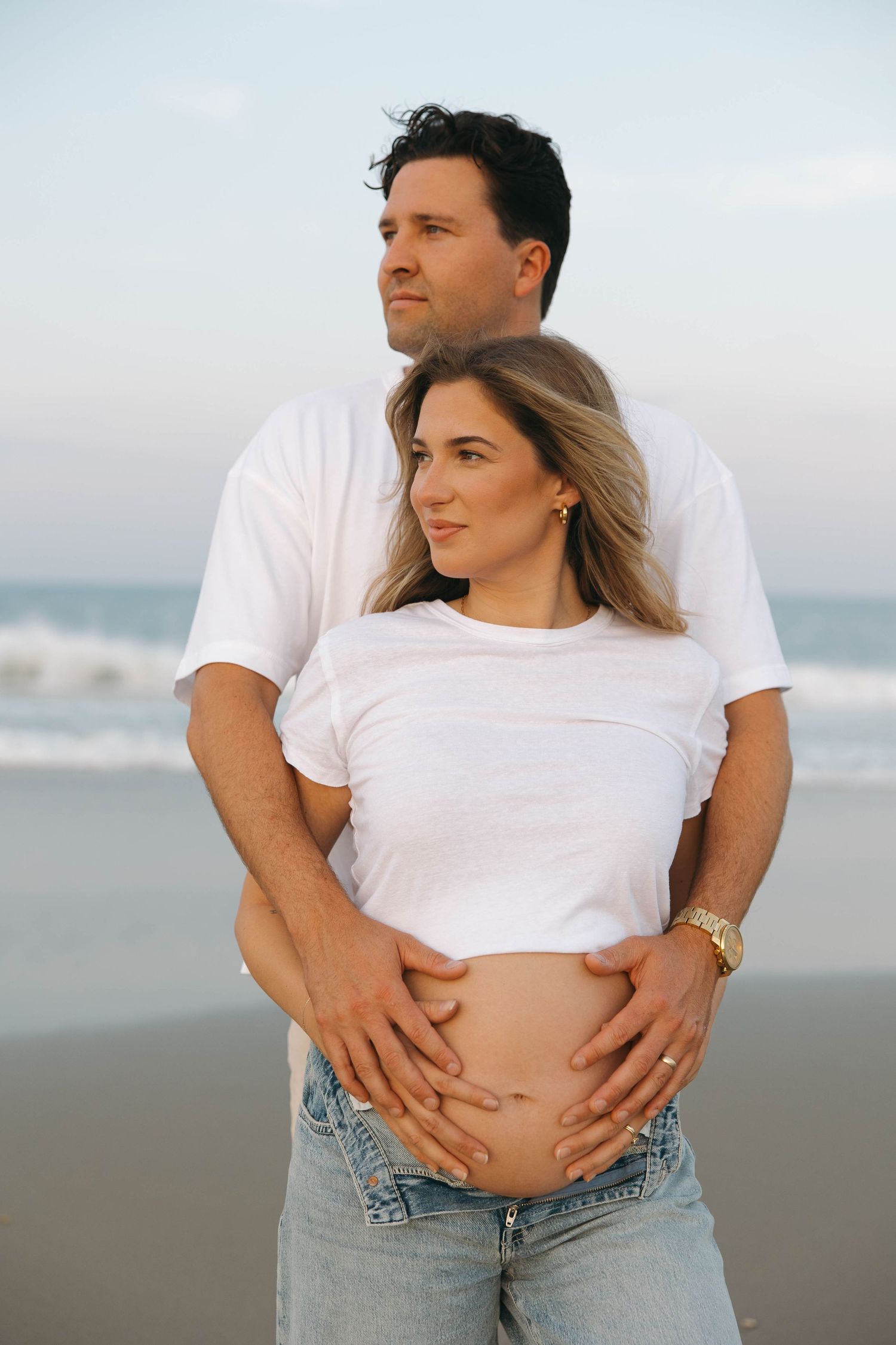 A couple in white shirts and jeans embraces on a misty beach during a maternity photoshoot.