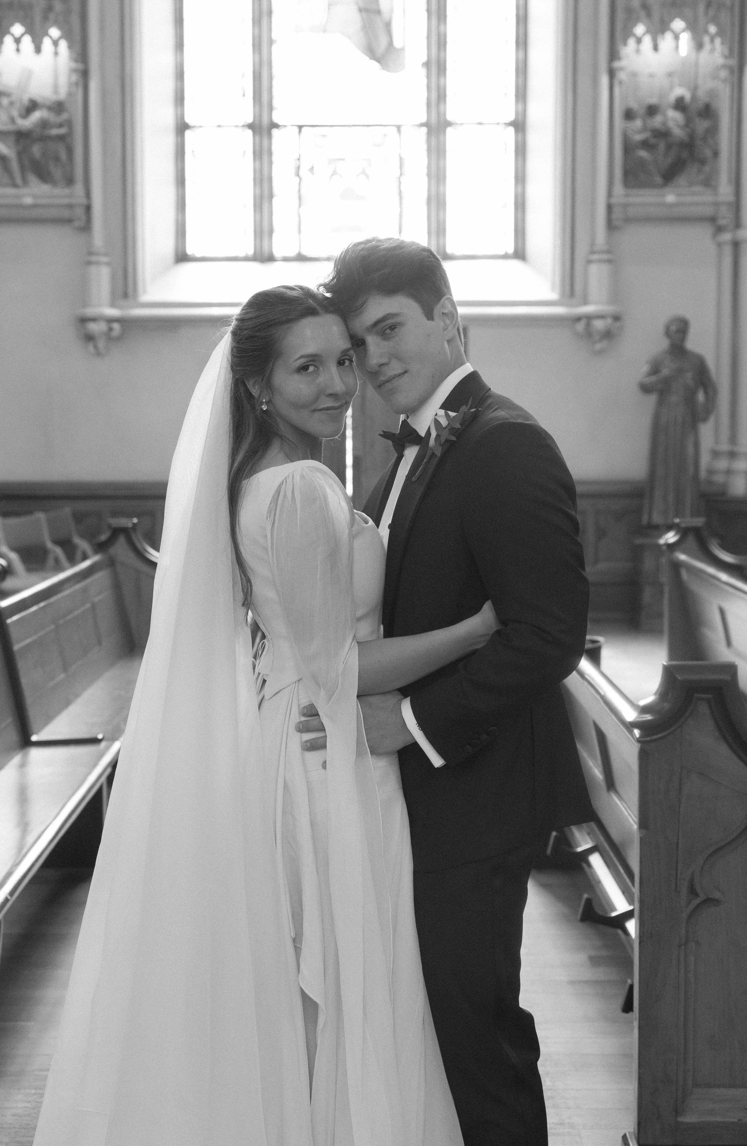 Black and white portrait of bride and groom embracing in church aisle with statues and pews behind.