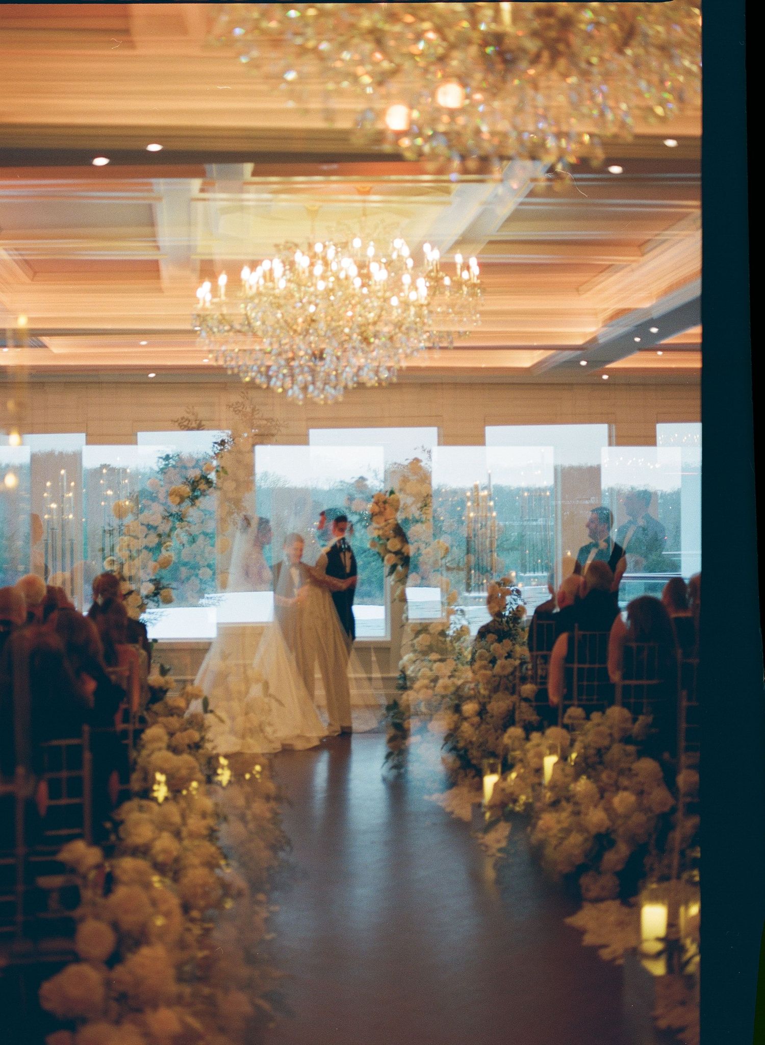 A wedding ceremony in a luxurious ballroom with crystal chandeliers and elegant floral arrangements lining the aisle.