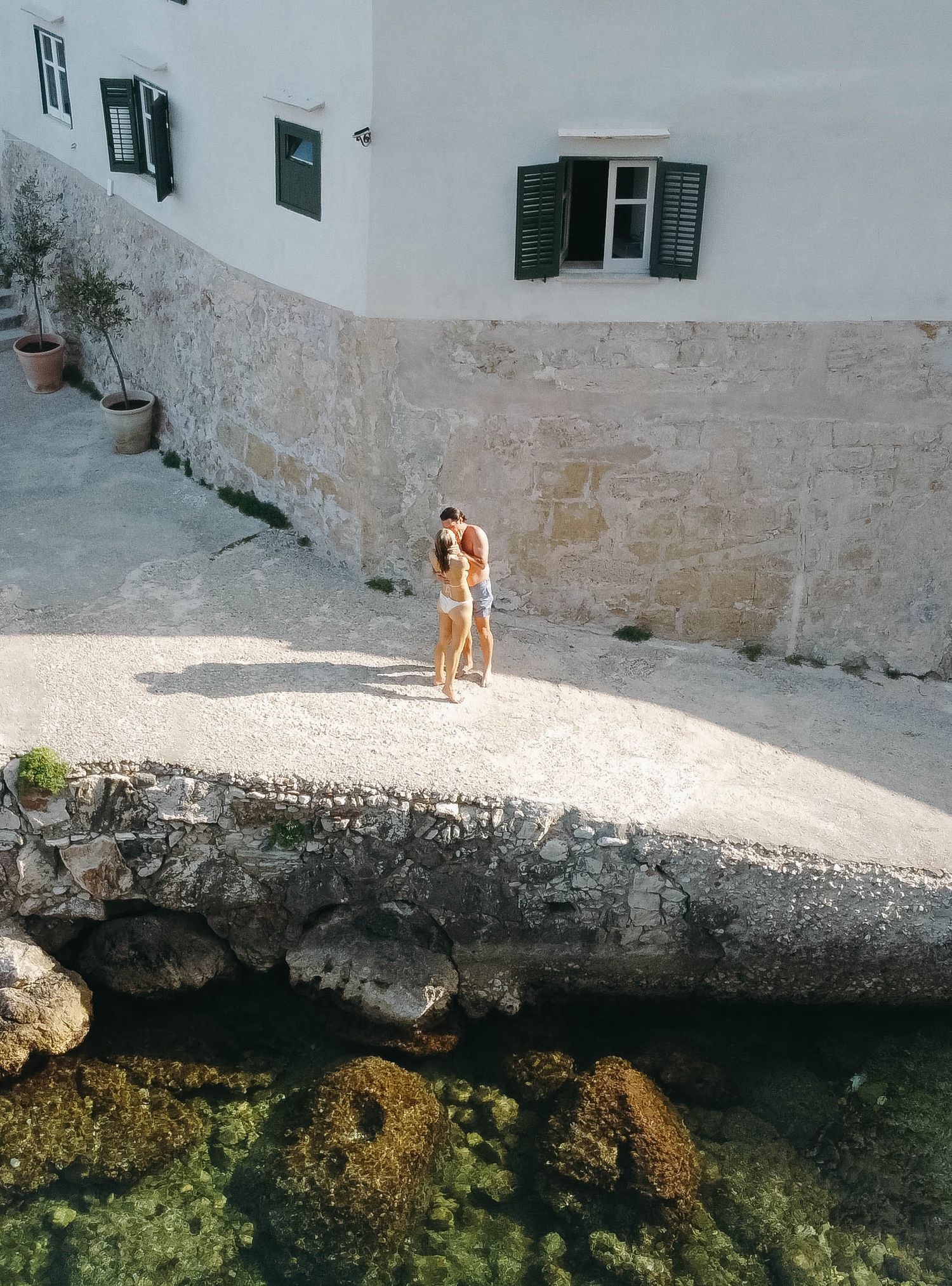 White stone terrace overlooking clear Mediterranean waters with moss-covered rocks below.