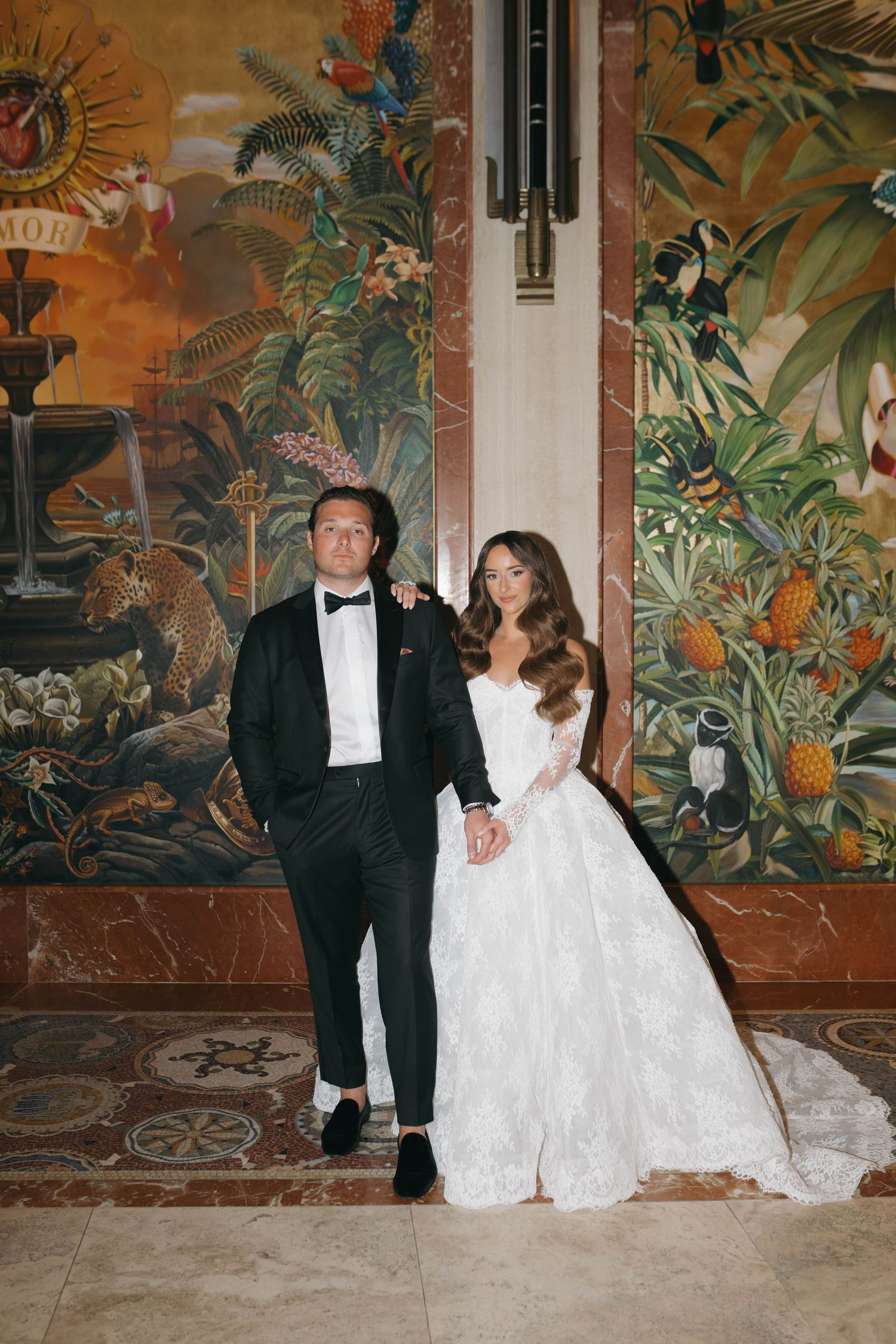 Wedding couple poses against a vibrant tropical mural wall in formal attire.