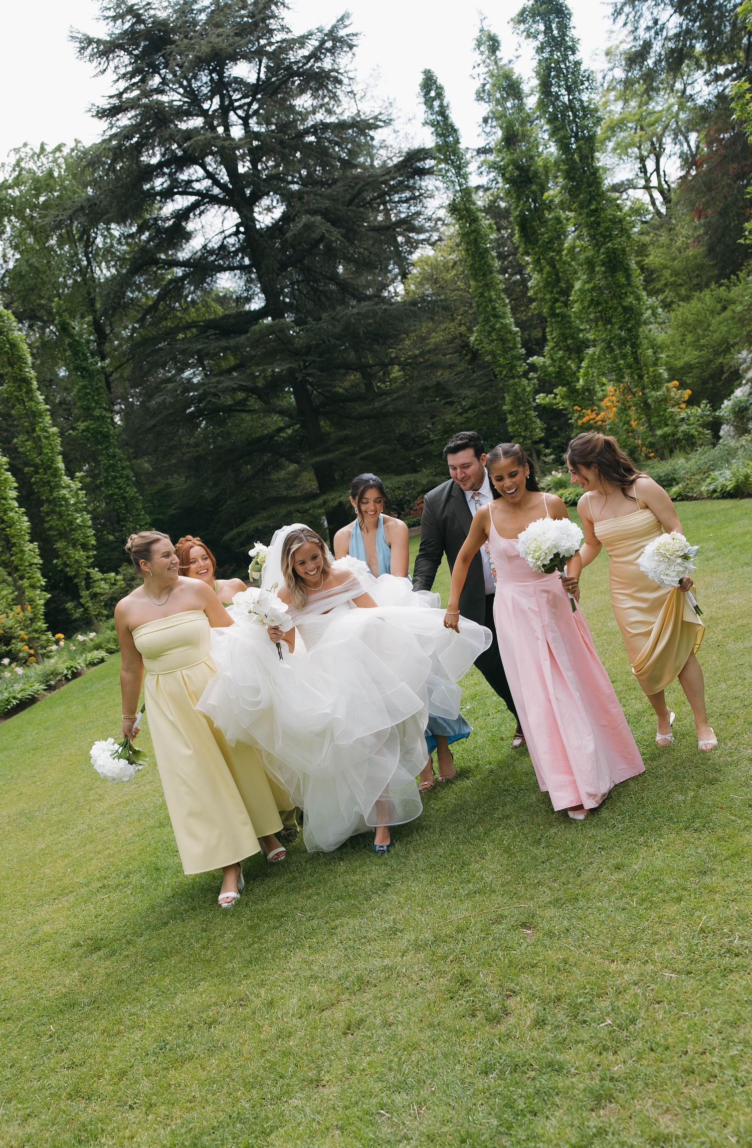 Bridesmaids in pastel dresses running playfully across a green lawn.