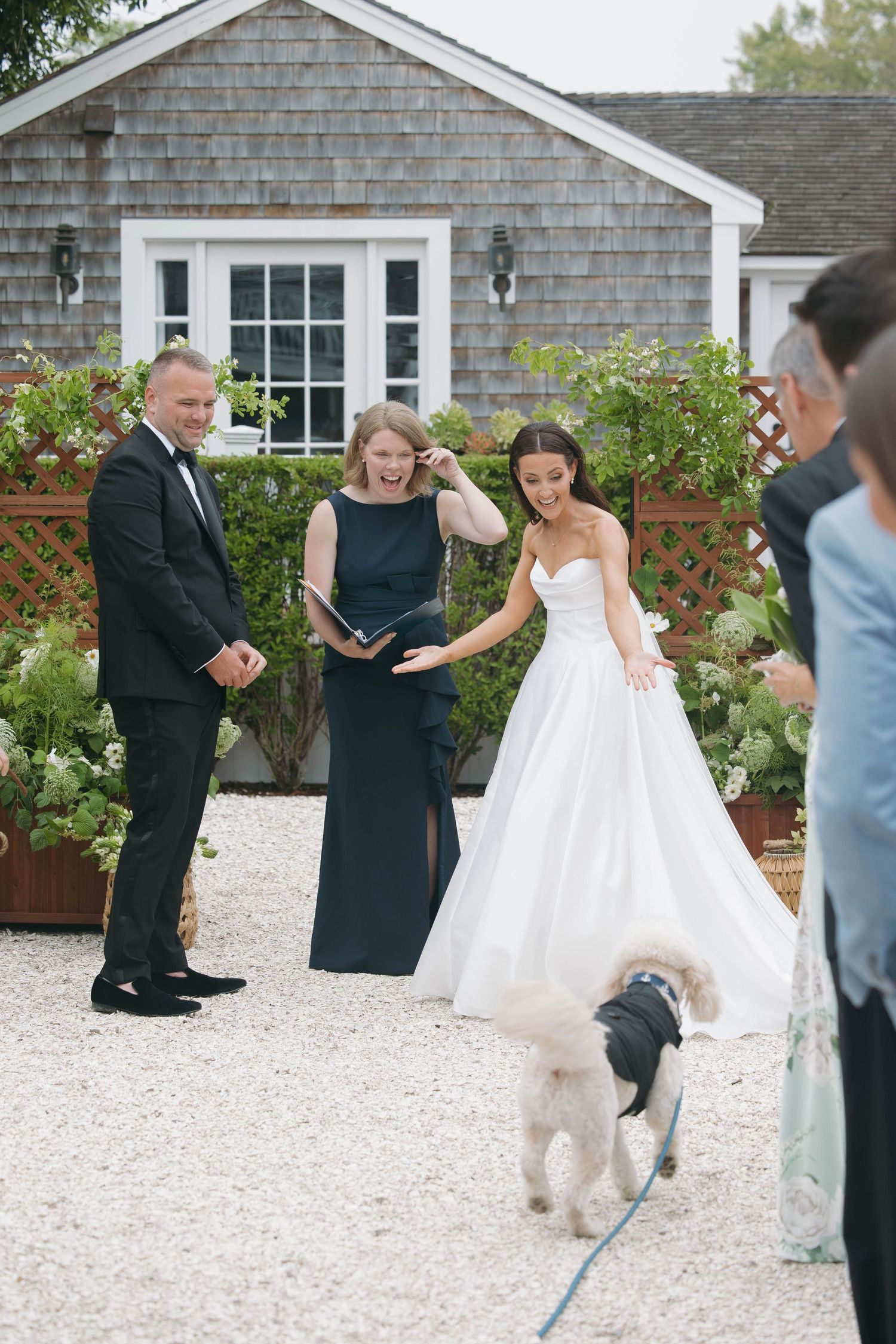 Wedding ceremony participants walk with a small white dog on a gravel path by a shingled building.