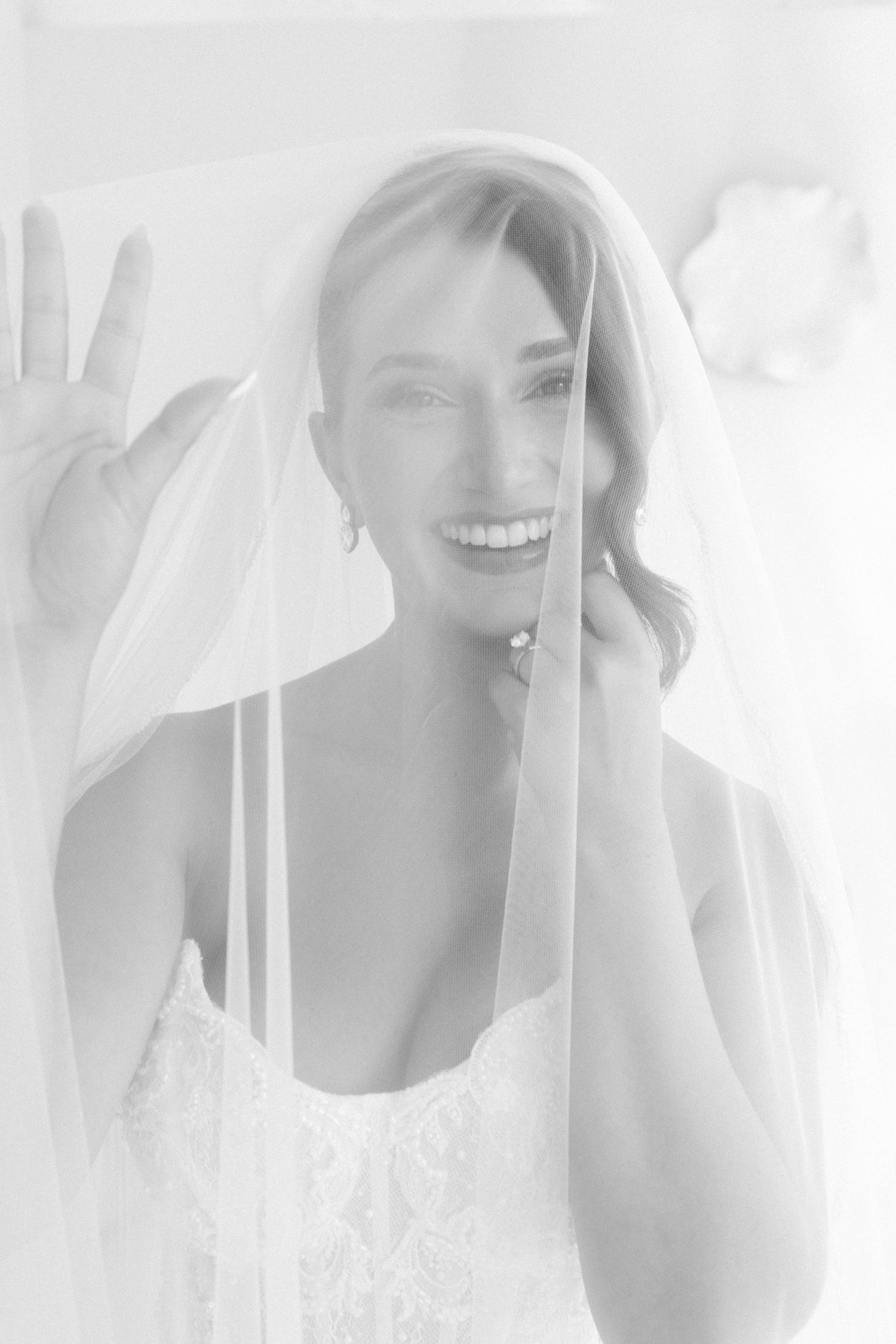 Black and white portrait of a smiling bride holding her veil, wearing a lace wedding dress and delicate earrings.