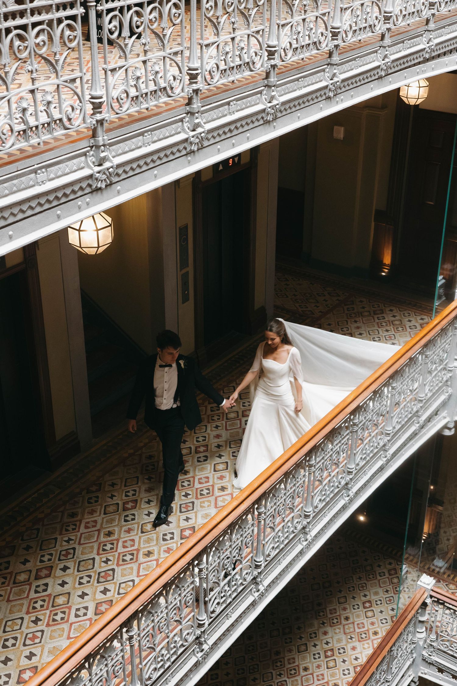 Bride and groom walk hand-in-hand along ornate balcony railing, wedding dress train trailing behind in elegant historic venue.