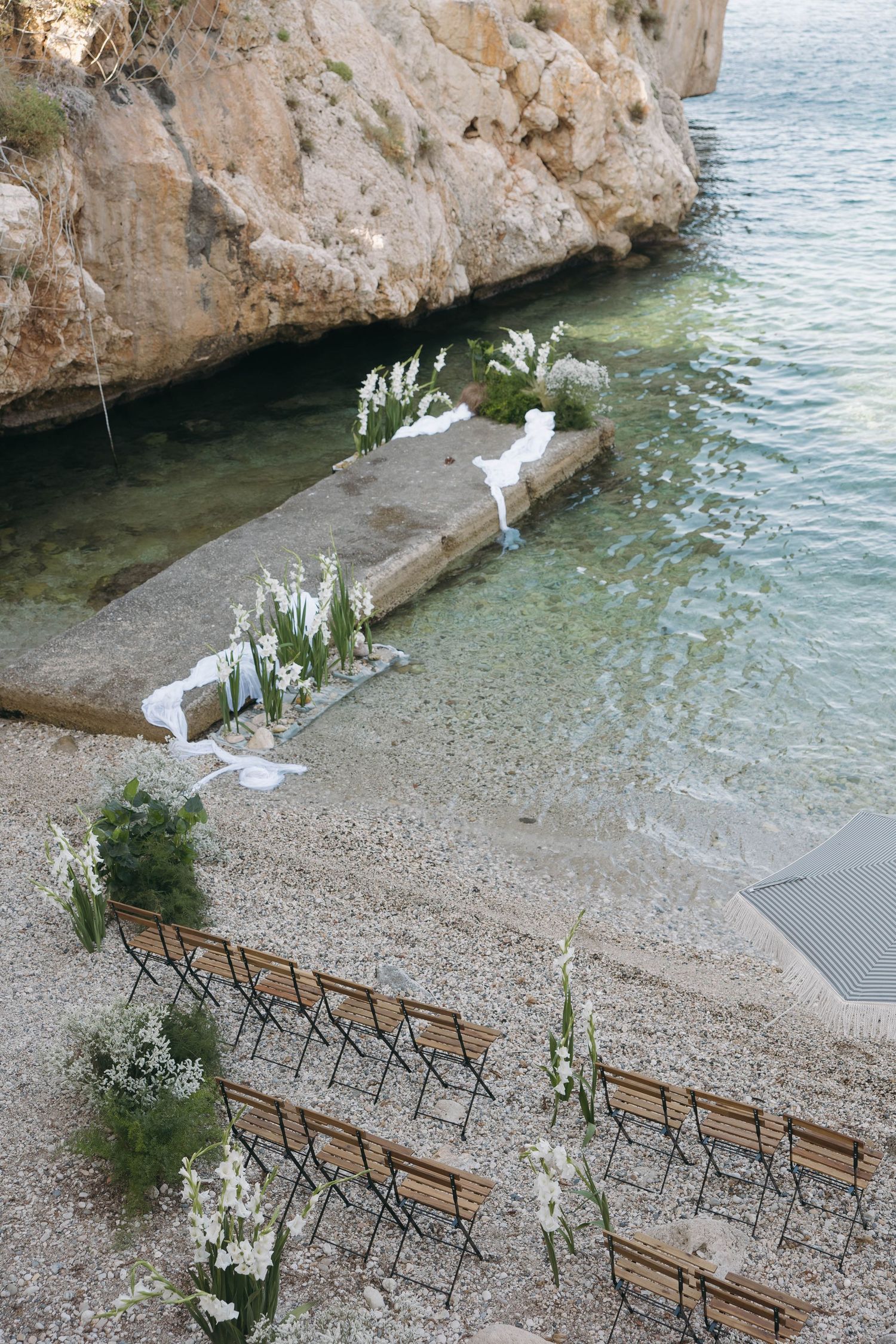 Coastal wedding ceremony site with white floral arrangements and wooden seating by crystal clear waters.