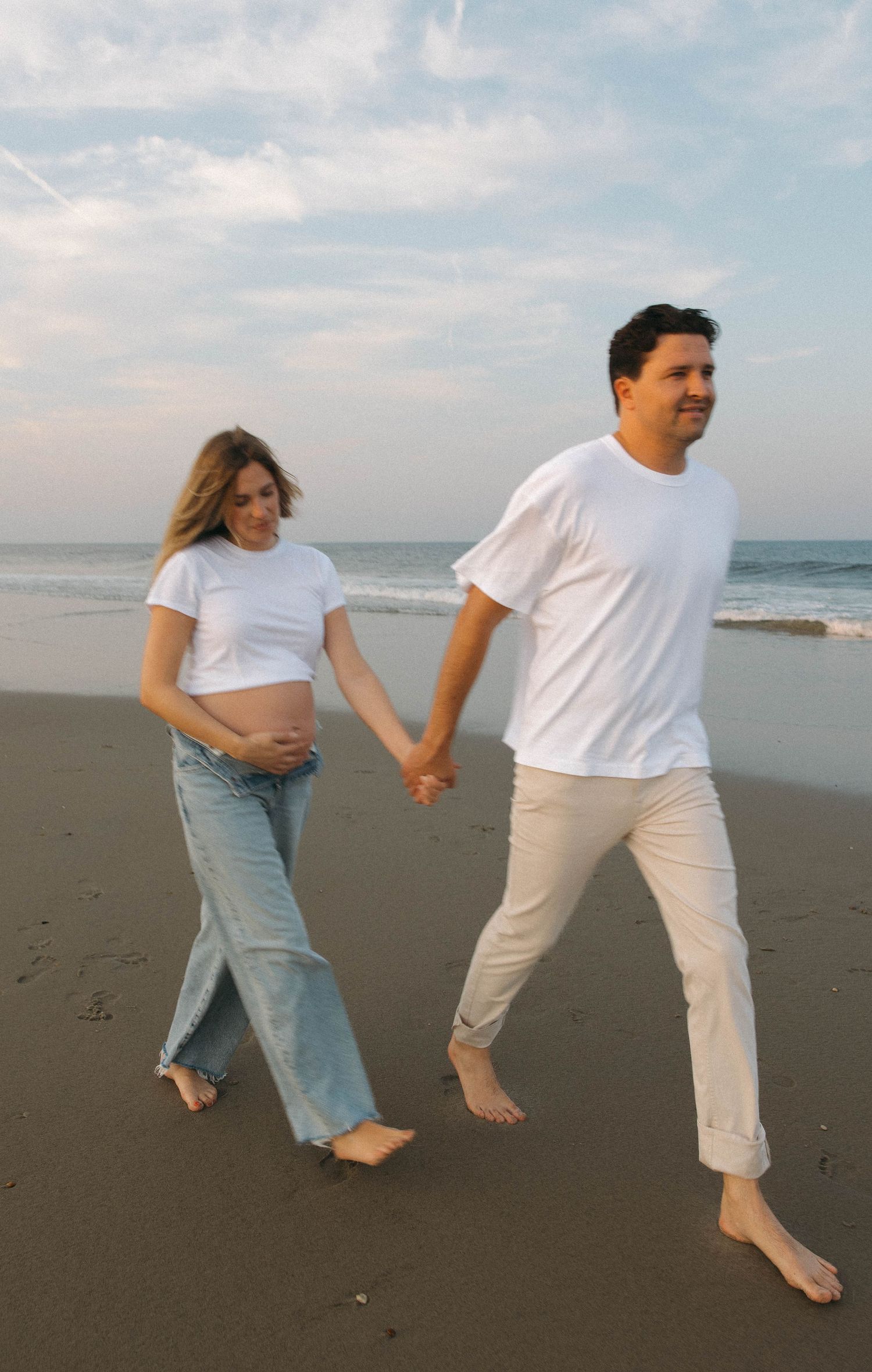 A couple in white and denim outfits playfully walking and holding hands on a scenic beach at sunset.