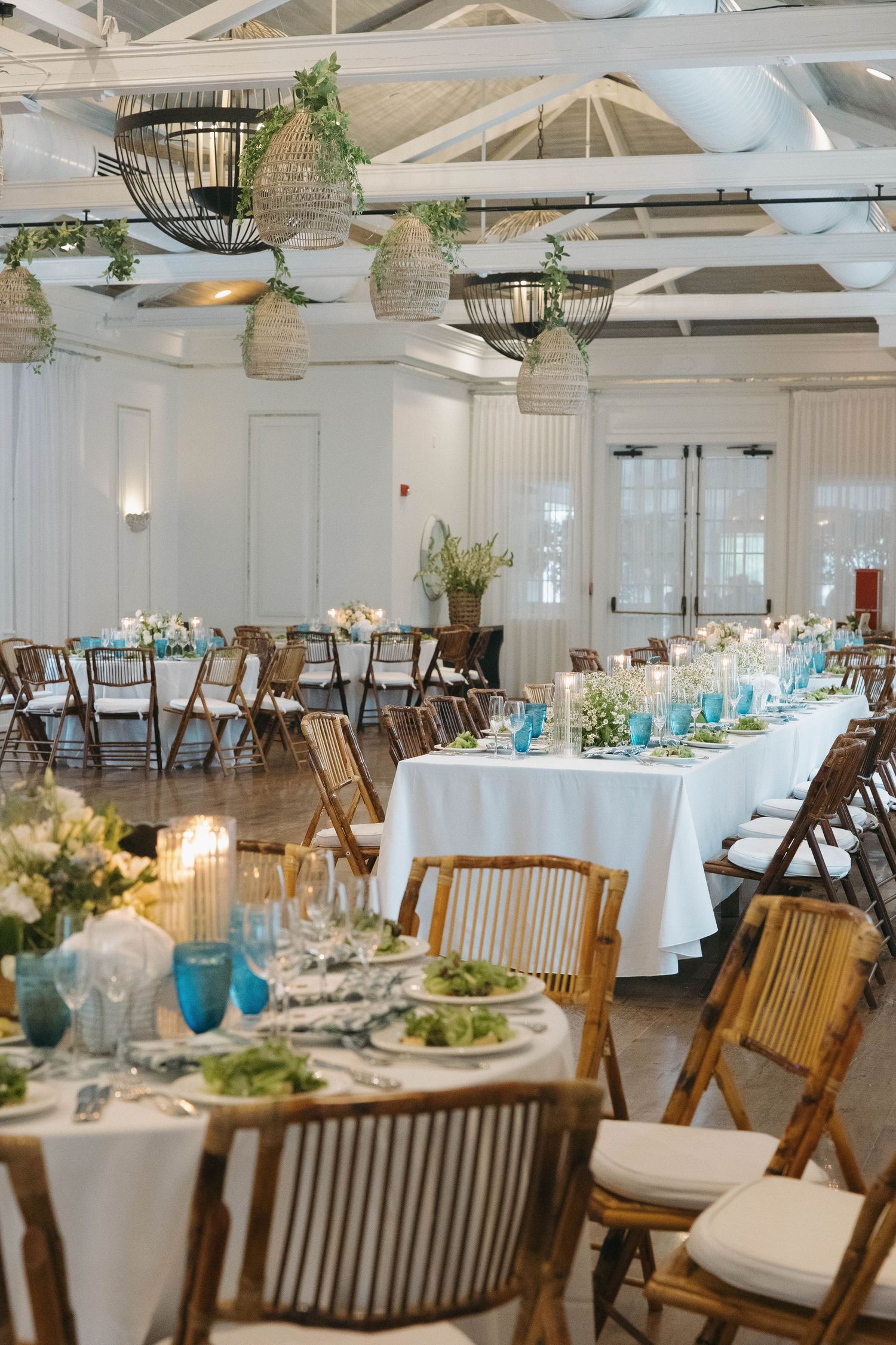 Wedding reception space with rustic wooden chairs and white tablecloths in an airy white venue.