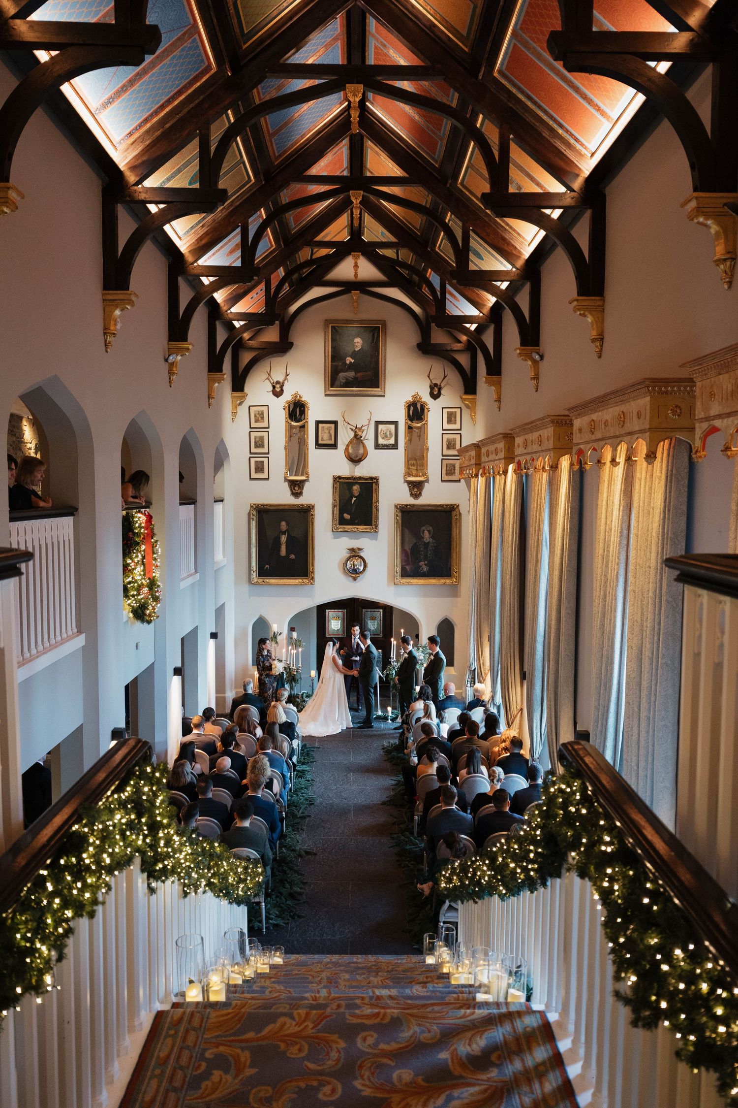 A grand castle interior with vaulted wooden beams and columns decorated with holiday lights.