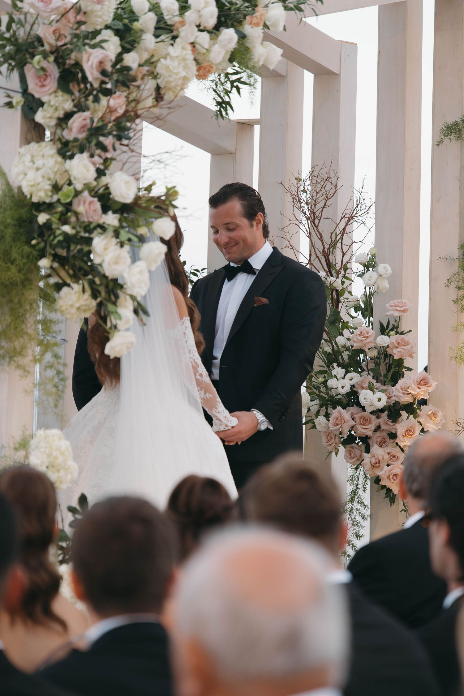 Wedding ceremony in progress under a white pergola with abundant floral decorations and seated guests.
