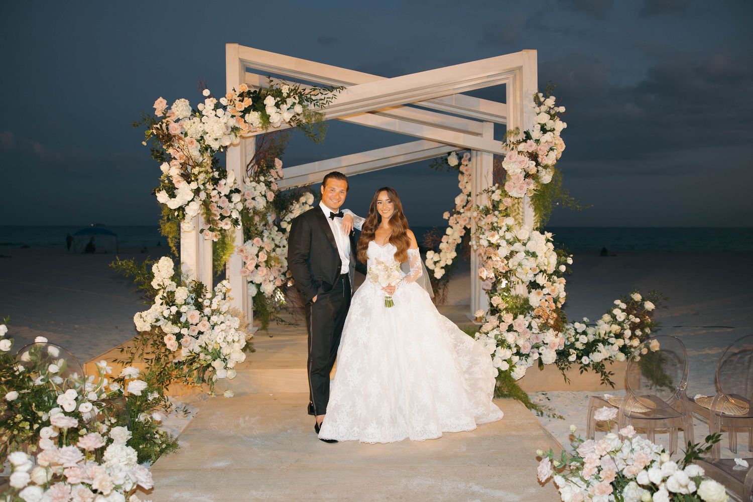 Elegant beach wedding ceremony with floral-adorned pergola against a dusky ocean backdrop.
