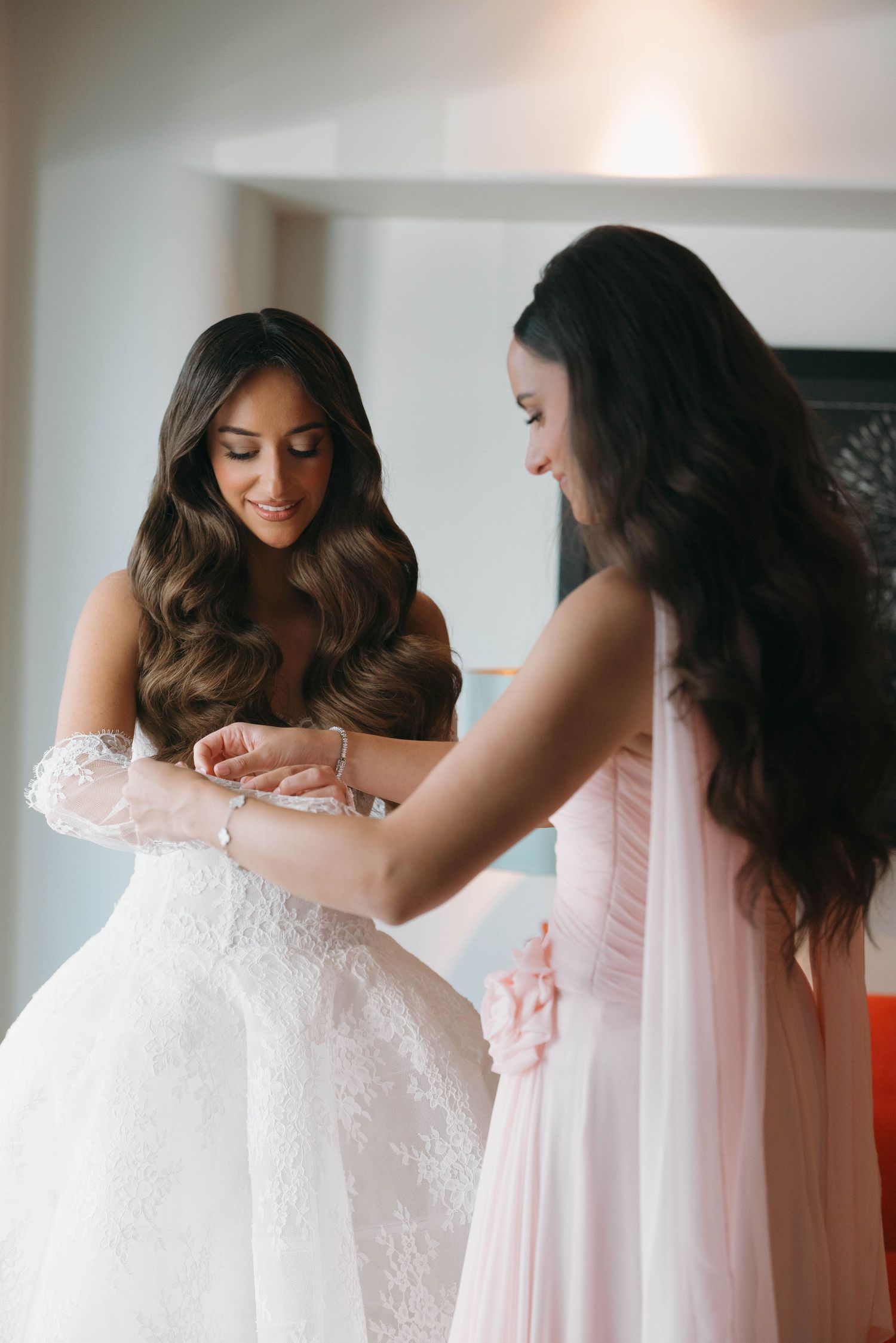 Person adjusting white lace wedding dress with assistance in sunlit room.