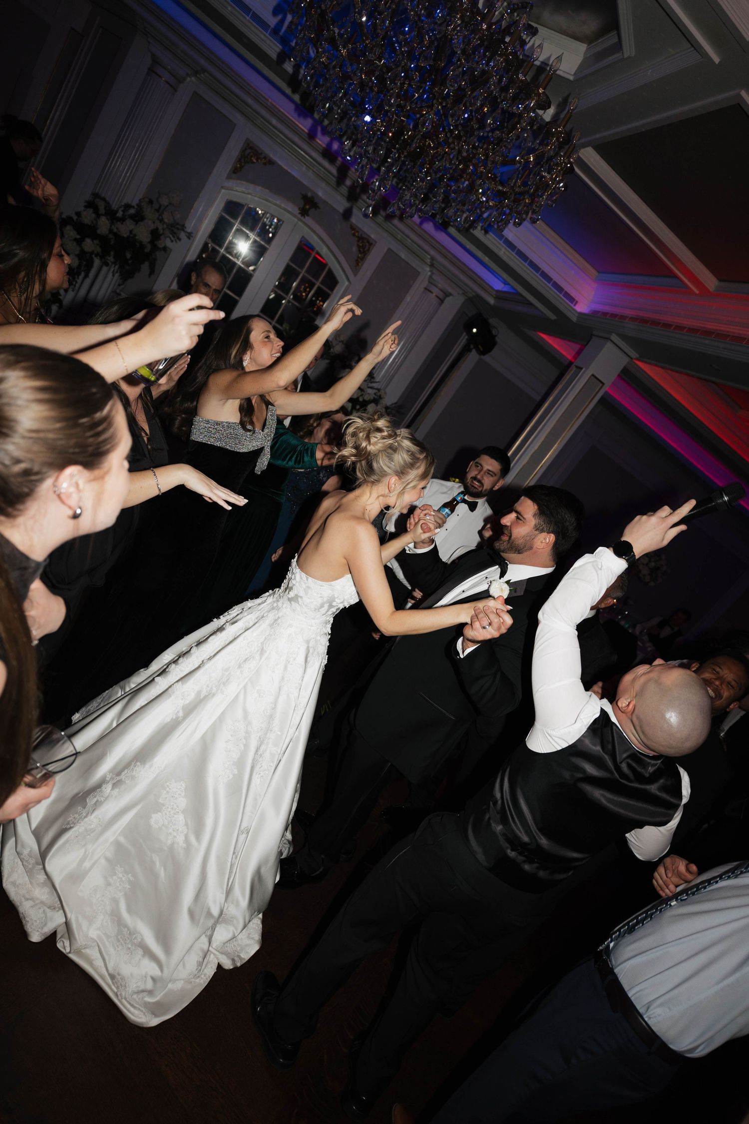 Wedding guests dance and celebrate with dramatic lighting and chandelier overhead.